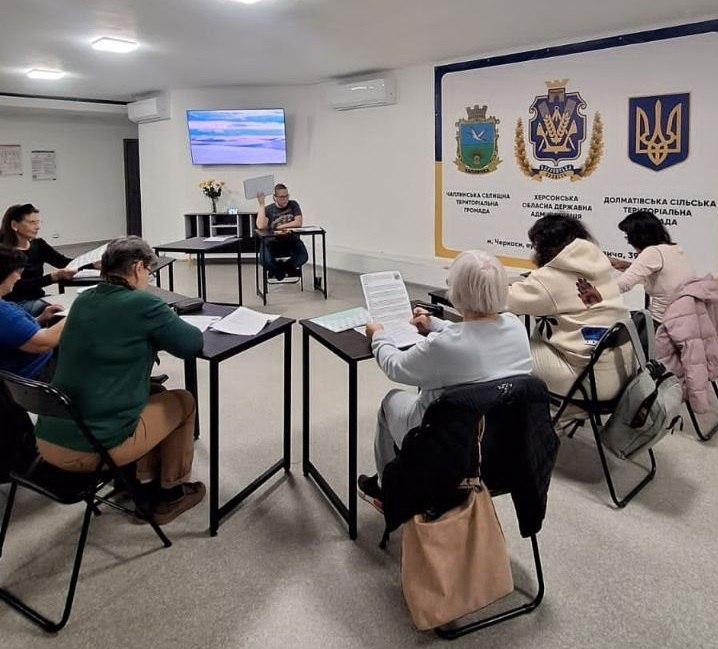 Група жінок-переселенок із Херсонщини вдосконалює навички спілкування під час заняття з англійської мови в координаційному центрі «Вільні разом» у Черкасах, де шість учасниць сидять за чотирма окремими чорними металевими столами, розставленими півколом, та опрацьовують роздруковані навчальні матеріали й бланки з інтерактивними вправами, а перед ними в торці кімнати за столом сидить викладач у чорній футболці та окулярах, який підіймає вгору аркуш паперу для демонстрації завдання, ліворуч від нього на стіні встановлено ввімкнений плазмовий екран із пейзажем, на передньому плані зліва сидить жінка в зеленому светрі, справа — жінка у світлому одязі з великою бежевою сумкою на стільці, тоді як на стіні праворуч розміщено великий офіційний білий стенд із Державним Гербом України, гербами громад та написами «Чаплинська селищна територіальна громада», «Херсонська обласна державна адміністрація» та «Долматівська сільська територіальна громада».