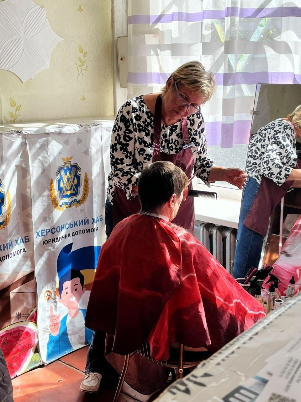 Жінка-перукарка у блузці з чорно-білим анімалістичним принтом та бордовому захисному фартуху робить стрижку відвідувачці, яка сидить перед нею у червоному перукарському пеньюарі під час надання безоплатних перукарських послуг для внутрішньо переміщених осіб із Херсонщини в координаційному центрі «Вільні разом» у Кривому Розі, де майстриня акуратно підстригає волосся клієнтки за допомогою ножиць та гребінця, а ліворуч робоче місце облаштоване великим офіційним білим банером із золотим гербом міста Херсона та написом «ХЕРСОНСЬКИЙ ХАБ ЮРИДИЧНА ДОПОМОГА», під яким розміщено малюнок хлопчика на тлі синьо-жовтого прапора, водночас праворуч у великому вертикальному дзеркалі відображається сам процес роботи перукарки, а на столику поруч розставлені професійні спреї, флакони та перукарські інструменти біля великого вікна з бузковими шторами.