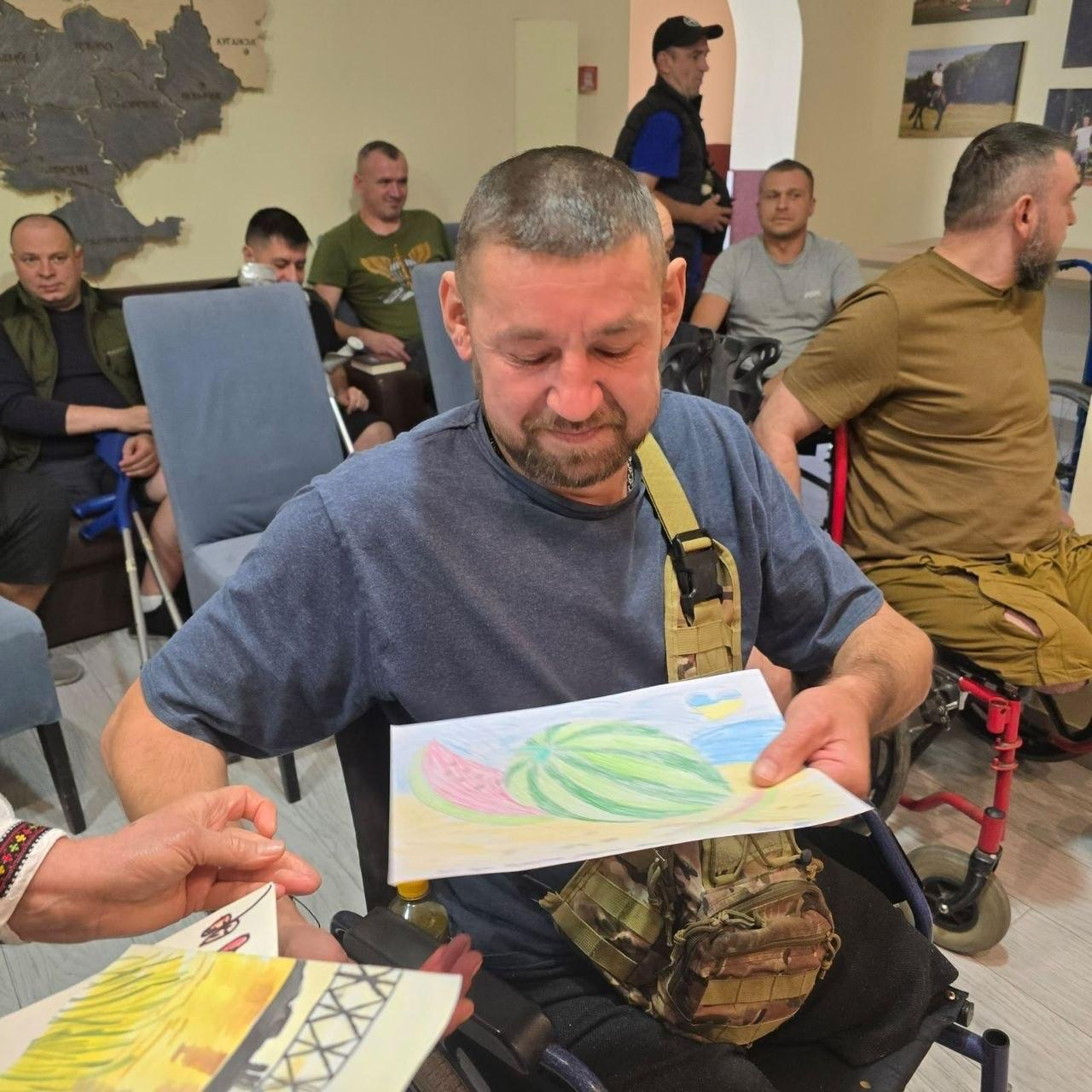 Український захисник, який проходить лікування та пересувається на кріслі колісному, з теплою посмішкою тримає в руках кольоровий дитячий малюнок із зображенням стиглого херсонського кавуна, отриманий під час візиту представників координаційного центру «Вільні разом» у Вінниці до лікарні для підтримки поранених воїнів, де боєць одягнений у синю футболку, через його плече закріплено тактичну військову сумку захисного кольору, перед ним зліва співробітниця хабу в білій вишиванці з червоно-чорним орнаментом тримає в руках інші дитячі листи та листівочки для передачі, водночас на задньому плані в залі медичного закладу на стільцях сидять інші поранені військовослужбовці та ветерани, які уважно спостерігають за подією, праворуч зафіксовано ще одного захисника у футболці кольору койот на кріслі колісному, а на світлій стіні кімнати ліворуч висить велика настінна карта України.