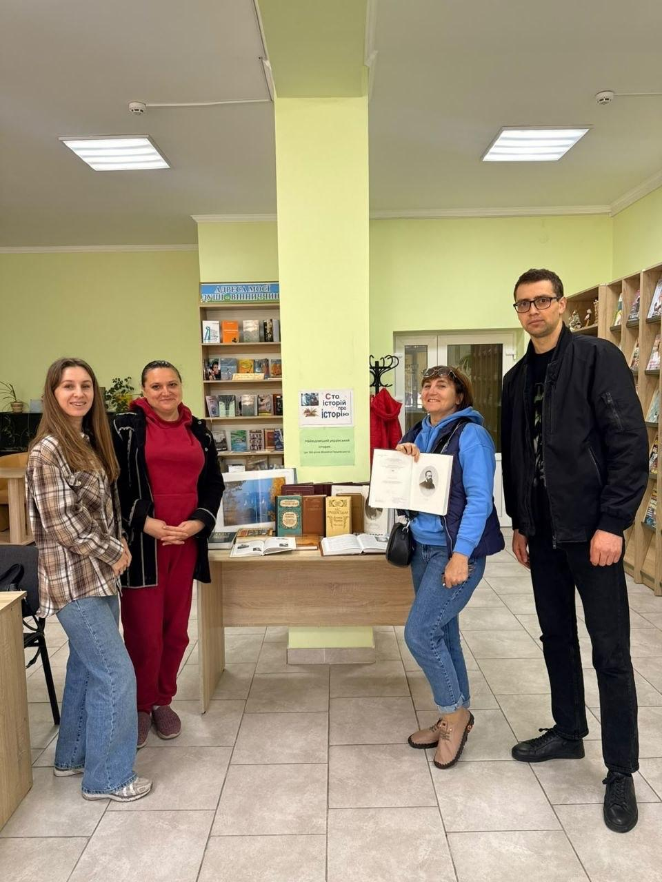 Чотири учасники заходу — представники координаційного центру «Вільні разом» у Вінниці та переселенці з Херсонщини — позують для спільного фото під час відвідин книжкової виставки «Найвідоміший український історик», організованої у бібліотеці до 160-річчя Михайла Грушевського, де у світлому залі з ніжно-зеленими стінами та великою колоною по центру вони вишикувалися навколо дерев'яного столу з розгорнутими історичними книгами, художніми репродукціями й портретами в рамках, причому ліворуч стоїть дівчина у картатій сорочці та блакитних джинсах поруч із жінкою в бордовому спортивному костюмі та темному кардигані, водночас праворуч від столу жінка у синьому худі, жилетці та джинсах тримає в руках відкриту книгу з портретом видатного історика, посміхаючись у кадр разом із високим чоловіком у чорній куртці, чорній футболці з малюнком, темних штанах та окулярах, тоді як на задньому плані кадру видно великі книжкові шафи, стелажі з літературою, плакат із написом «Сто історій про історію» та скляні міжкімнатні двері.