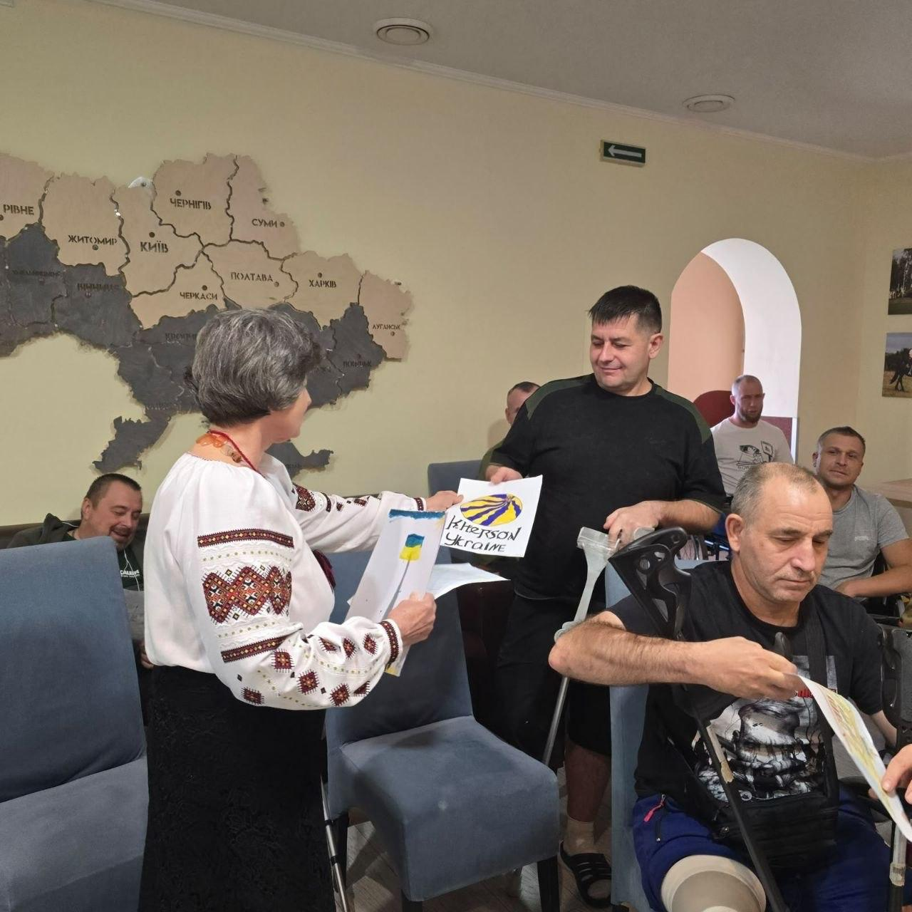 Співробітниця координаційного центру «Вільні разом» у Вінниці в білій вишиванці з традиційним червоно-чорним орнаментом передає патріотичні дитячі листи та малюнки пораненим українським захисникам під час візиту до лікарні для підтримки воїнів, де ліворуч жінка похилого віку з коротким сивим волоссям тримає в руках малюнок синьо-жовтого прапора та інші роботи юних переселенців із Херсонщини, а по центру кадру поранений боєць у чорній футболці, спираючись на милиці, з посмішкою приймає аркуш із намальованим кавуновим сонцем та написом «Kherson Ukraine», водночас праворуч на передньому плані сидить інший військовослужбовець, який також уважно розглядає отримане привітання, тримаючи милицю під пахвою, а на задньому плані в залі медичного закладу на стільцях сидітимуть інші пацієнти, які проходять лікування, тоді як на світлій стіні кімнати ліворуч розміщено велику карту України з назвами областей.