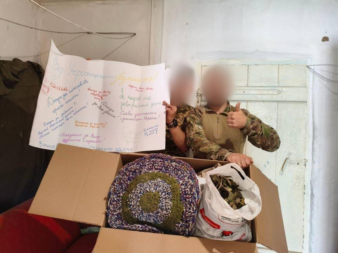 Двоє українських військовослужбовців у камуфляжній формі тримають велику картонну коробку з отриманою гуманітарною допомогою, надісланою переселенцями з Херсонщини в межах акції «Від Херсонщини - з вдячністю» від координаційного центру «Вільні разом» у Вінниці, де на передньому плані всередині коробки видно великі круглі в'язані килимки ручної роботи та білий пластиковий пакет із завантаженими речами, причому боєць праворуч піднімає вгору великий палець на знак подяки, а за ними воїни розгорнули та тримають великий білий плакат-ватман, на якому різнокольоровими маркерами написані патріотичні гасла, слова підтримки й побажання від херсонців, зокрема «Від Херсонщини — з вдячністю!», «Дякую за захист!!!», «Слава захисникам і захисницям України!», «Віримо!» та «ЧЕКАЄМО ВДОМА!», водночас самі захисники зафіксовані з розмитими з міркувань безпеки обличчями в бліндажі чи приміщенні на тлі світлих дерев'яних дверей та стін.