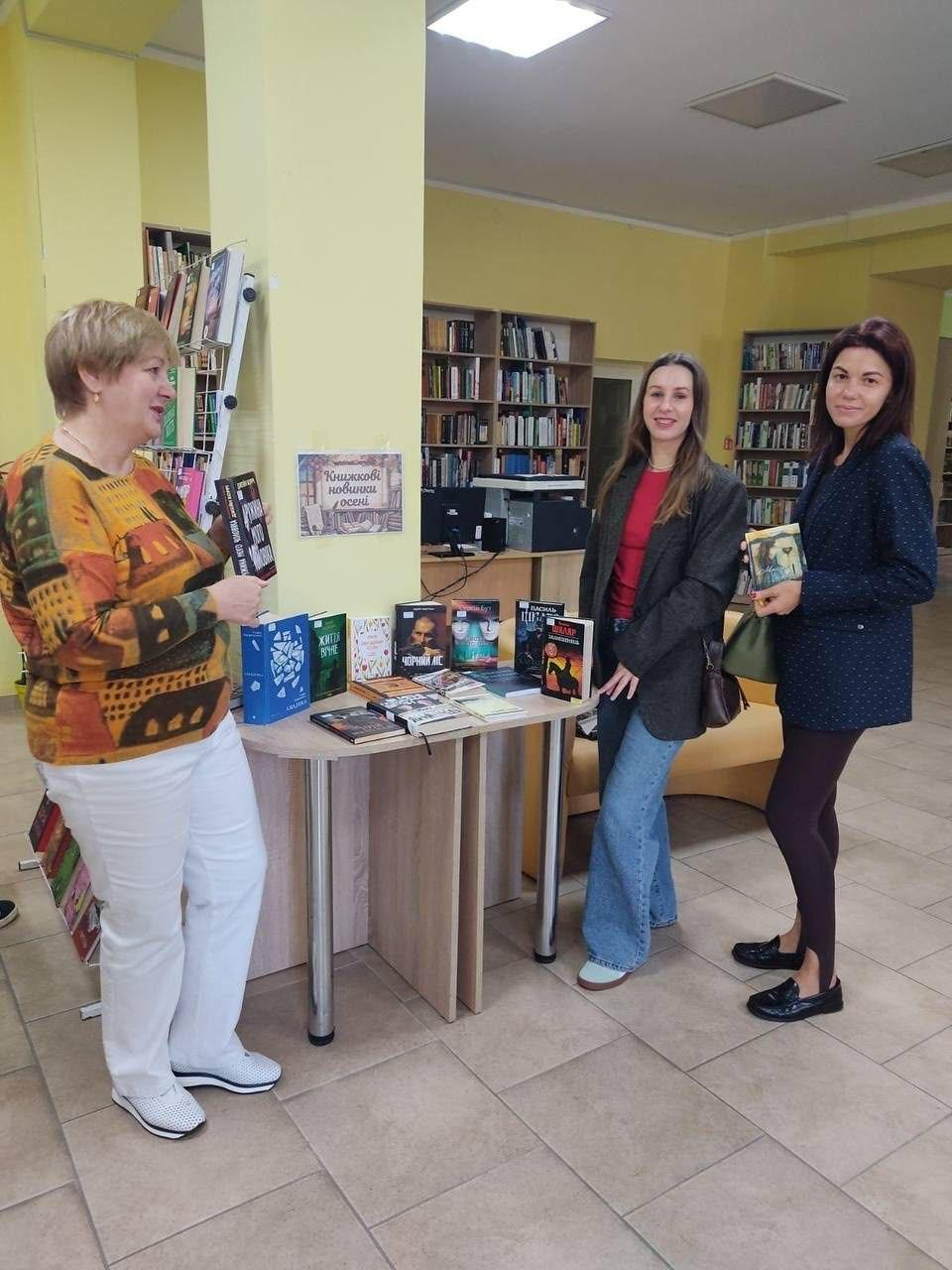 Три жінки — представниці координаційного центру «Вільні разом» у Вінниці та переселенки з Херсонщини — позують біля виставкового столу під час відвідин виставки «Книжкові новинки осені» у бібліотеці-філії №13 Вінницької міської ЦБС до Всеукраїнського дня бібліотек, де ліворуч стоїть співробітниця установи в яскравому помаранчево-жовтому светрі з малюнками та білих штанах, тримаючи в руках книгу і спілкуючись із відвідувачками, водночас праворуч по центру кадру стоїть дівчина у коричневому піджаку, червоній кофті та блакитних широких джинсах, а крайня праворуч жінка в темно-синьому подовженому піджаку в цяточку та темних лосинах тримає художню книгу в руках, причому всі вони зафіксовані навколо півкруглого дерев'яного столу з розставленою сучасною літературою, над яким на великій колоні кабінету закріплено малюнок-афішу заходу, а на задньому плані просторого залу видно високі книжкові шафи-стелажі з книгами та робоче місце бібліотекаря з комп'ютером.