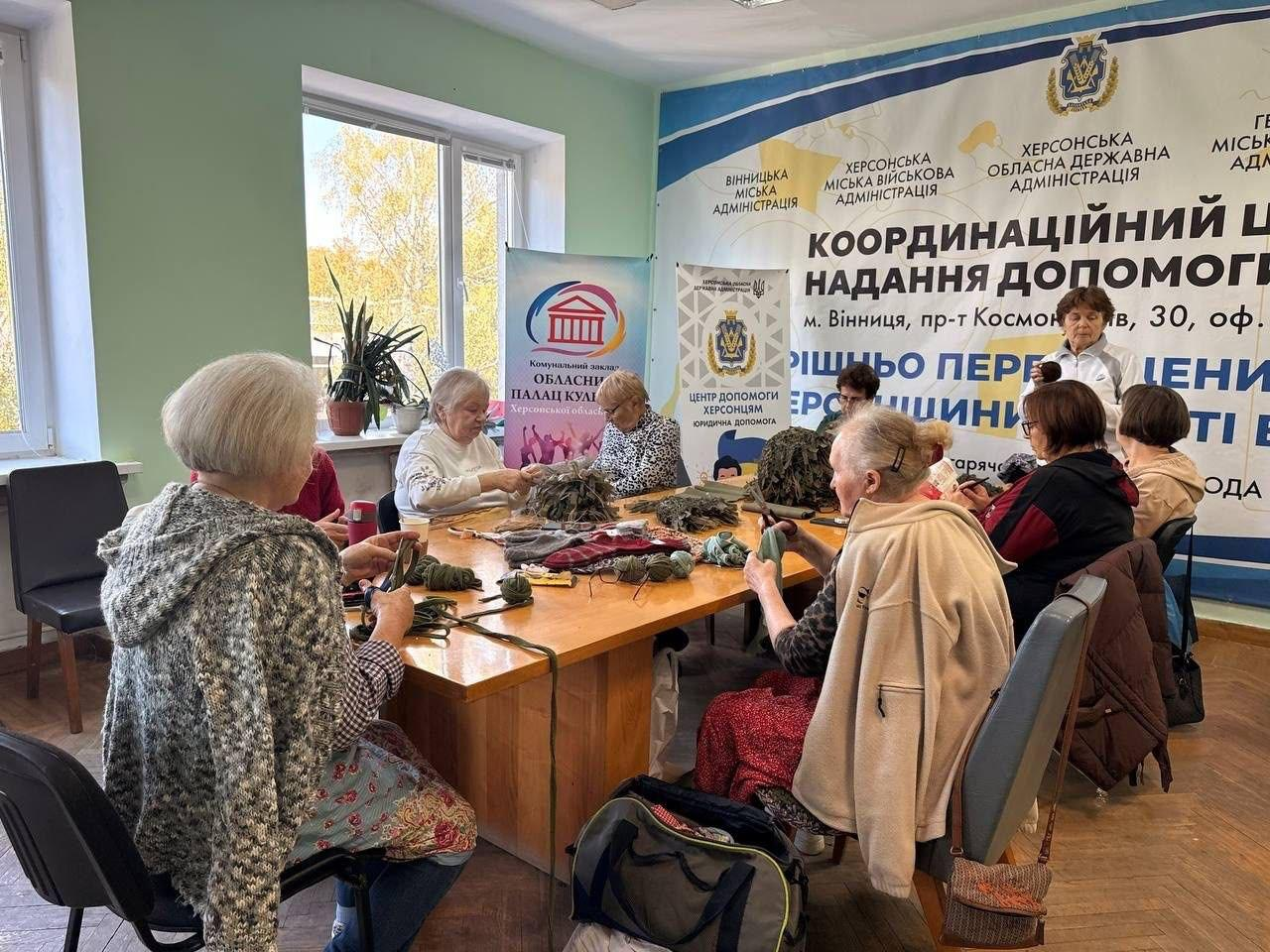 Велика група жінок-переселенок із Херсонщини займається плетінням нашоломників та теплих шкарпеток для ЗСУ під час благодійної патріотичної акції в координаційному центрі «Вільні разом» у Вінниці, де дев'ять учасниць різного віку сидять навколо великого дерев'яного столу у світлому кабінеті хабу й активно працюють із різнокольоровою вовняною пряжею, нитками та заготовками для плетіння, розкладеними по центру столу разом із ножицями, великим термосом та особистими речами, за якими на задньому плані встановлено великий офіційний білий банер із гербами та великими написами «КООРДИНАЦІЙНИЙ ЦЕНТР НАДАННЯ ДОПОМОГИ ВНУТРІШНЬО ПЕРЕМІЩЕНИМ ОСОБАМ ХЕРСОНЩИНИ», «Вінницька міська адміністрація», «Херсонська міська військова адміністрація» та «Херсонська обласна державна адміністрація», водночас ліворуч біля великого вікна з кімнатними рослинами розміщено ролап-стенд комунального закладу «Обласний Палац культури» Херсонської обласної ради, а біля столу на передньому плані лежить тактичний рюкзак кольору хакі.