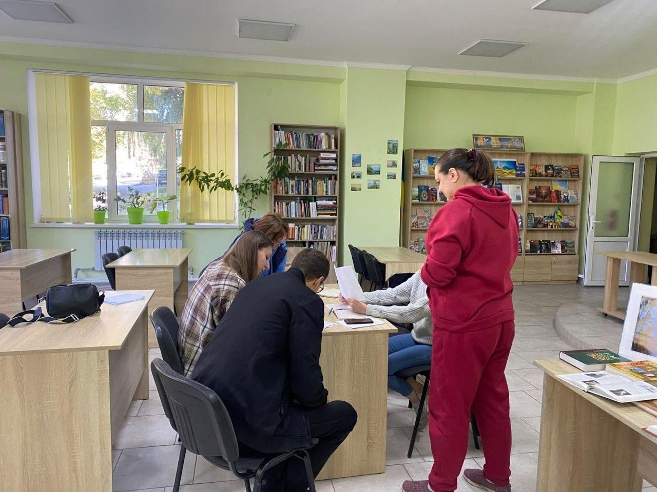 Група переселенців із Херсонської області отримує консультації з різних питань у просторому залі координаційного центру «Вільні разом» у Вінниці, де за робочим столом по центру кімнати сидять троє відвідувачів та фахівчиня хабу, яка переглядає надані документи й папери, водночас поруч із ними стоїть жінка в бордовому спортивному костюмі, а сам кабінет облаштований численними дерев'яними столами, стільцями та великими книжковими шафами з літературою на задньому плані, ліворуч розташоване велике світле вікно з кімнатними рослинами у зелених горщиках на підвіконні, а праворуч на окремому столі розкладено книги та художні репродукції.