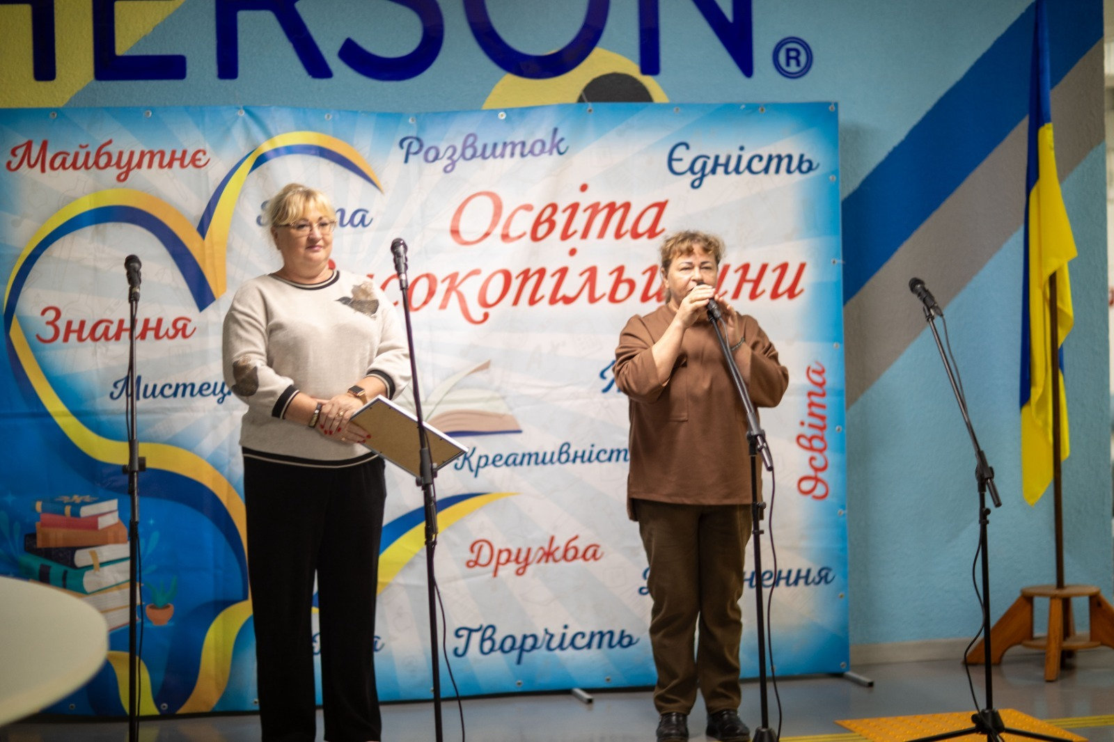 Дві жінки виступають на сцені перед мікрофонами під час урочистостей та концерту до Дня працівників освіти у Високопільській громаді, де ліворуч стоїть представниця керівництва Високопільської селищної військової адміністрації у світлій кофті та чорних штанах, яка тримає в руках офіційну папку з документами для привітання, а праворуч інша промовця у коричневій блузці та темних штанах виступає з промовою, причому обидві жінки зафіксовані на тлі великого яскравого брендованого банера з малюнками книг, серця та великими написами «Освіта Високопільщини», «Майбутнє», «Розвиток», «Єдність», «Знання», «Дружба», «Творчість», водночас у правій частині сцени на дерев'яній підставці встановлено Державний прапор України.