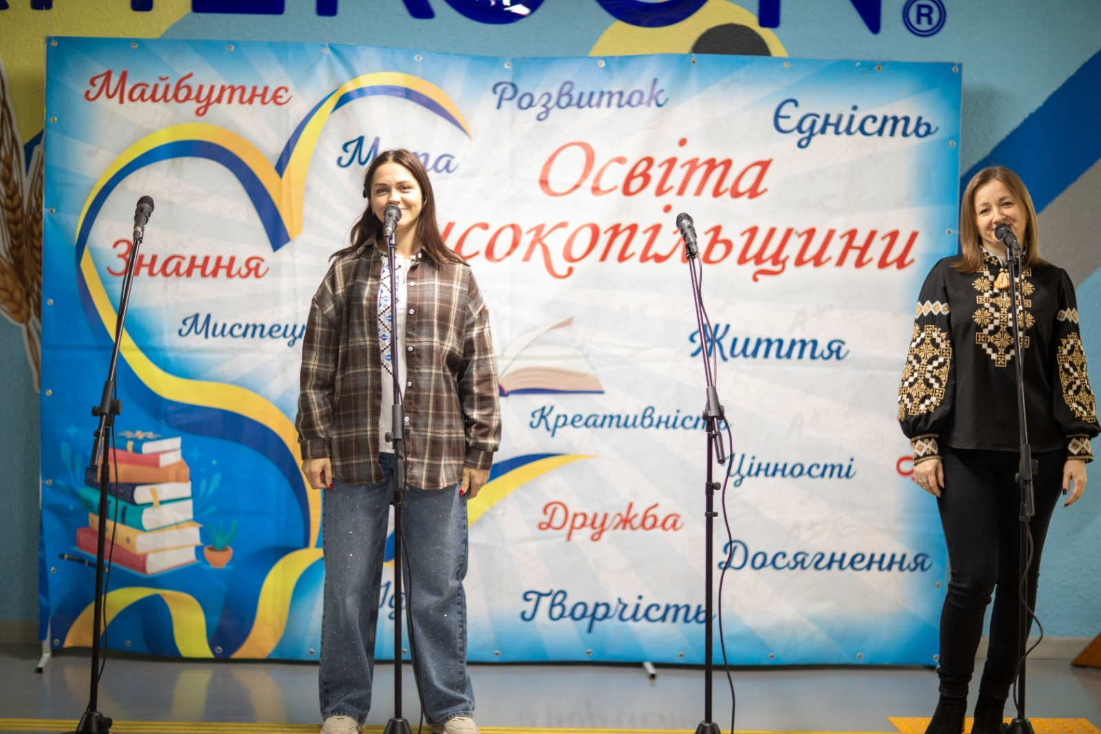 Дві жінки виступають на сцені перед мікрофонами під час святкового концерту до Дня працівників освіти у Високопільській громаді, де ліворуч стоїть дівчина у коричневій картатій сорочці поверх світлої футболки з традиційним орнаментом та у широких блакитних джинсах, яка з посмішкою співає або промовляє у мікрофон, а праворуч інша учасниця заходу в чорній вишиванці з багатим золотаво-білим геометричним візерунком на рукавах та грудях і чорних облягаючих штанях також посміхається в кадр, перебуваючи на тлі великого офіційного блакитно-білого банера з малюнками розгорнутих книг, жовто-синього серця та великими написами «Освіта Високопільщини», «Майбутнє», «Знання», «Мистецтво», «Розвиток», «Єдність», «Життя», «Цінності», «Досягнення», «Дружба» та «Творчість».