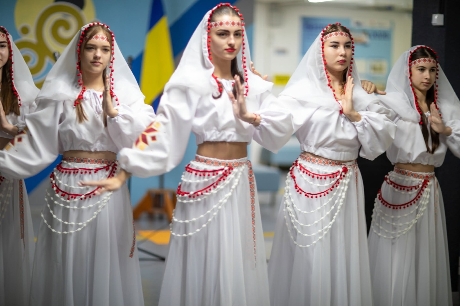 Юні учасниці місцевого творчого колективу у стилізованих білих українських костюмах виконують хореографічний номер під час святкового концерту до Дня працівників освіти у Високопільській громаді, де дівчата вишикувалися в один ряд на сцені, будучи одягненими в короткі білі блузи-топи з вишитими геометричними візерунками на широких рукавах та довгі легкі білі спідниці, прикрашені на поясі червоно-білими намистами, багатошаровими стрічками та декоративними помпонами, водночас на головах танцівниць закріплені білі намітки-хустки з червоною окантовкою та патріотичними пов'язками, а на задньому плані сцени ліворуч частково видно офіційний банер та встановлено Державний прапор України.