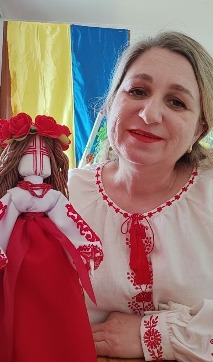 Майстриня та художня керівниця Олександрівського сільського будинку культури Каланчацької селищної ради Світлана Кудревська у красивій білій вишиванці з червоним орнаментом на грудях та пишних манжетах рукавів з посмішкою позує для фото поруч зі своєю автентичною авторською лялькою-мотанкою, з якою вона виборола диплом першого ступеня на міжнародному фестивалі мистецтв «Оксамитовий сезон» в Одесі. Творча робота майстрині представлена у вигляді традиційної української мотанки в червоній спідниці та білій вишитій блузі, з пишним вінком із червоних троянд на голові та вишитим червоним хрестом замість обличчя, а сама Світлана Кудревська зафіксована крупним планом на тлі великого синьо-жовтого Державного прапора України та прапора Херсонської області.