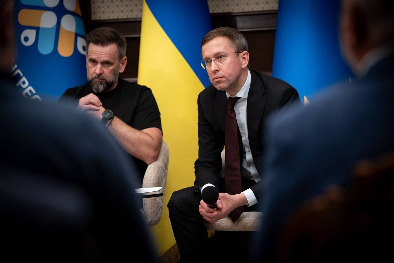Прем'єр-міністр України Сергій Корецький (праворуч, в окулярах, класичному чорному костюмі, білій сорочці та темно-бордовій краватці) сидить у кріслі з мікрофоном у руках та зосереджено виступає під час засідання Конгресу місцевих та регіональних влад при Президентові України, присвяченого обговоренню ситуації з державними фінансами. Ліворуч від нього в кріслі сидить заступник керівника Офісу Президента України та Голова Президії Конгресу Віктор Микита в чорній футболці, уважно слухаючи колегу, обоє посадовців перебувають на тлі великого синьо-жовтого Державного прапора України та іншого офіційного синього стяга з круговою емблемою заходу, де частково видно напис