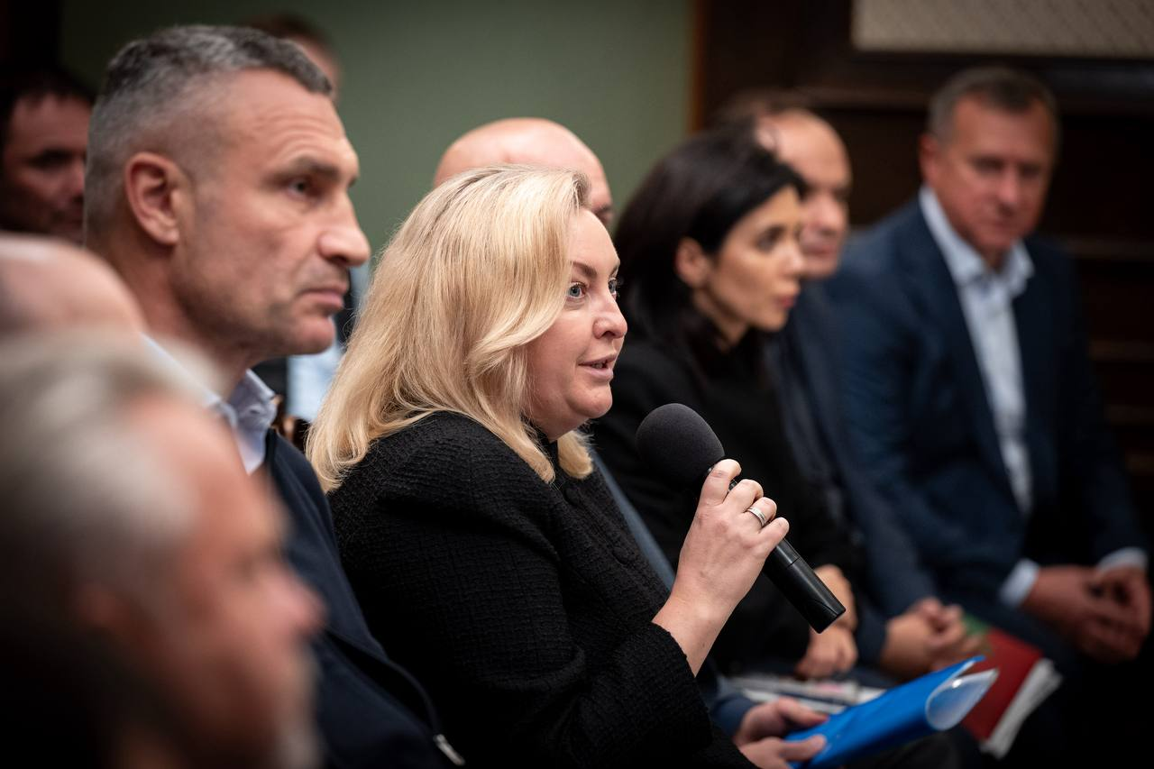 Учасниця засідання Конгресу місцевих та регіональних влад при Президентові України (по центру, зі світлим волоссям, у чорному жакеті) тримає в руці мікрофон і зосереджено виступає під час обговорення ситуації з державними фінансами та вимушеним режимом жорсткої економії. Ліворуч від неї сидить мер Києва Віталій Кличко у класичному костюмі та світлій сорочці, уважно слухаючи промову, поруч у ряд розмістилися інші присутні урядовці, голови міст та народні депутати, серед яких праворуч у профіль виділяється жінка з темним волоссям у чорному вбранні, яка тримає синю папку з документами, а на задньому плані кадру на тлі стіни видно чоловіків у ділових костюмах, тоді як обличчя людей на першому плані ліворуч залишаються розмитими.