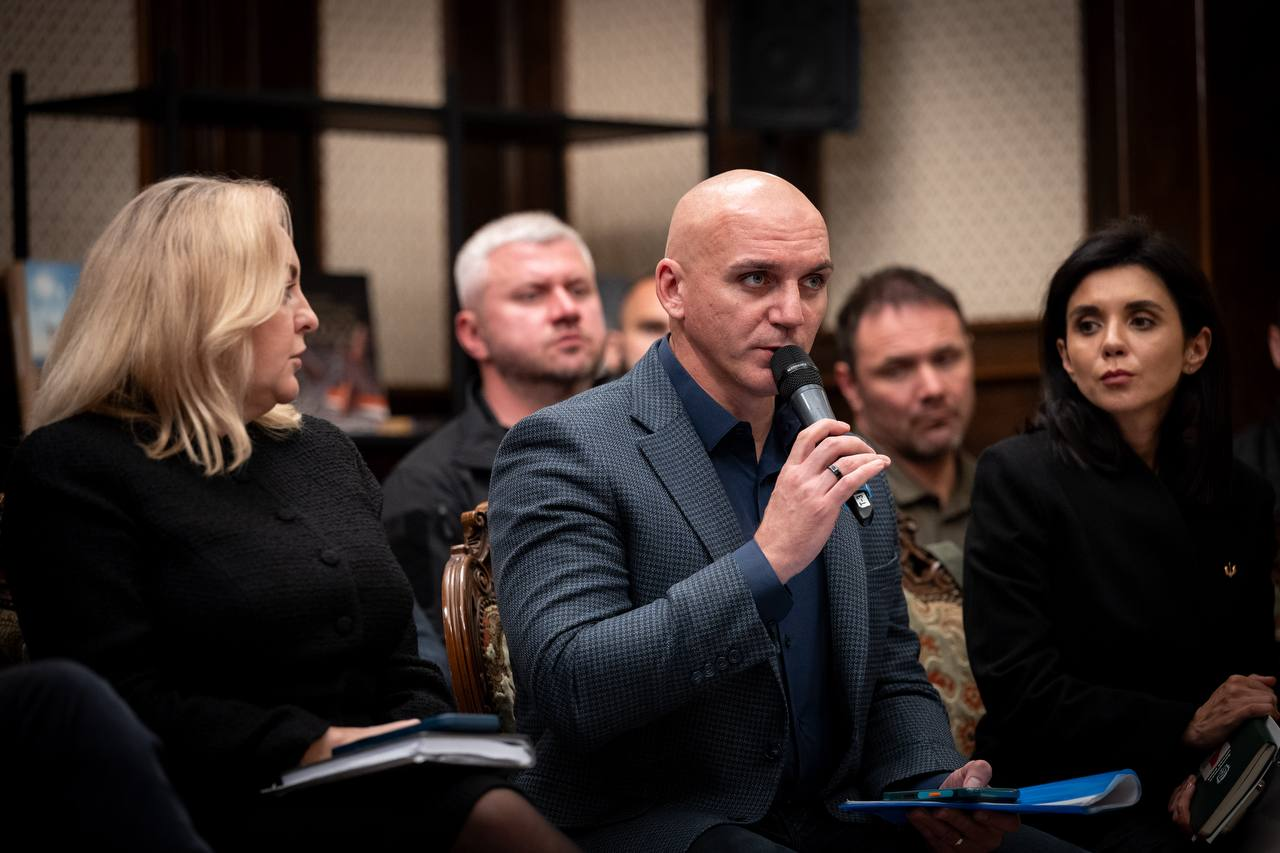 Миколаївський міський голова Олександр Сєнкевич (по центру, у синьому піджаку та темній сорочці) тримає в руці мікрофон і зосереджено виступає з конструктивними пропозиціями під час засідання Конгресу місцевих та регіональних влад при Президентові України, присвяченого обговоренню ситуації з державними фінансами. Ліворуч від нього сидить учасниця заходу зі світлим волоссям у чорному жакеті з відкритим блокнотом на колінах, яка уважно слухає промову, праворуч розмістилася жінка з темним волоссям у чорному вбранні з синьою папкою в руках, а на задньому плані кадру на тлі дерев'яних інтер'єрних панелей та стелажів видно інших учасників засідання, урядовців та народних депутатів, серед яких виділяється сивочолий чоловік у темному одязі.