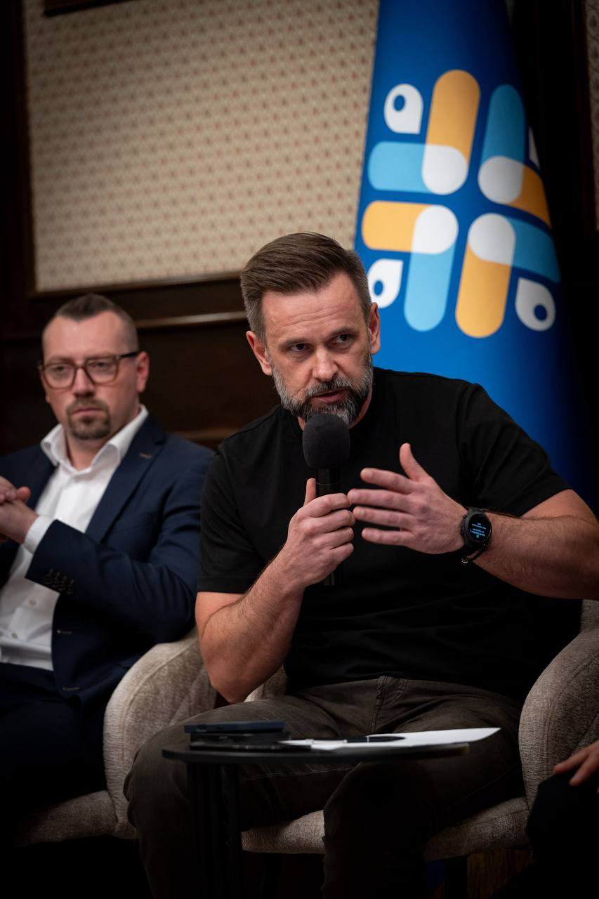 Міністр у справах громад, територій та внутрішньо переміщених осіб України Віталій Безгін (ліворуч, в окулярах, класичному синьому піджаку та білій сорочці) сидить у кріслі, склавши руки перед собою, та уважно слухає виступ заступника керівника Офісу Президента України Віктора Микити (по центру, в чорній футболці та темних штанах), який тримає в руці мікрофон і зосереджено виступає під час засідання Конгресу місцевих та регіональних влад при Президентові України, присвяченого обговоренню ситуації з державними фінансами, де праворуч на задньому плані встановлено великий офіційний синій стяг Конгресу із круговою різнокольоровою емблемою у жовтих, блакитних та білих тонах, а перед ними на маленькому круглому столику лежать документи й мобільний телефон.