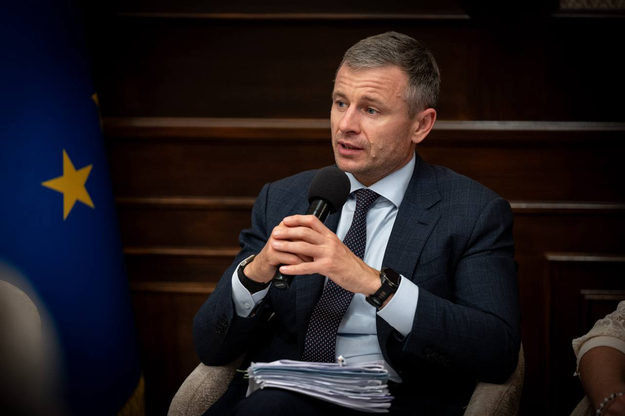 Міністр фінансів України Сергій Марченко (по центру, в картатому темно-синьому костюмі, світлій сорочці та темній краватці) сидить у кріслі з мікрофоном у руках і зосереджено виступає під час засідання Конгресу місцевих та регіональних влад при Президентові України, на колінах у нього лежить велика папка з робочими документами й роздрукованими матеріалами, ліворуч на задньому плані видно синій прапор Європейського Союзу із золотою зіркою, а в інтер'єрі залу домінують класичні дерев'яні стінові панелі.