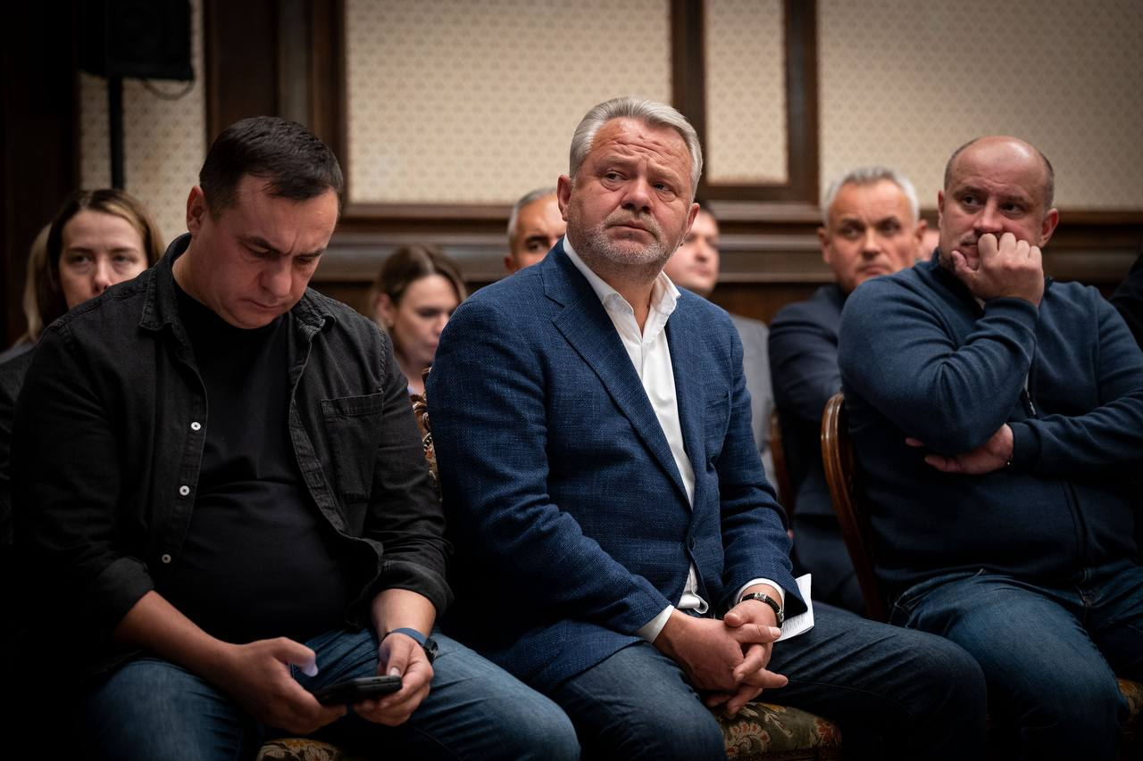 Учасники засідання Конгресу місцевих та регіональних влад при Президентові України сидять у ряд та уважно слухають виступи спікерів, де по центру кадру сидить сивочолий чоловік у синьому піджаку, білій сорочці та темних штанах, зчепивши пальці рук перед собою та зосереджено дивлячись у бік сцени. Ліворуч від нього сидить чоловік у чорній сорочці поверх чорної футболки та в синіх джинсах, який нахилив голову вниз та дивиться у свій мобільний телефон, а праворуч на стільці розмістився інший учасник заходу в темно-синій кофті на блискавці та джинсах, який підніс ліву руку до обличчя та підпер підборіддя, тоді як на задньому плані кадру на тлі світлих стін та дерев'яних панелей видно обличчя інших присутніх представників громад та урядовців.
