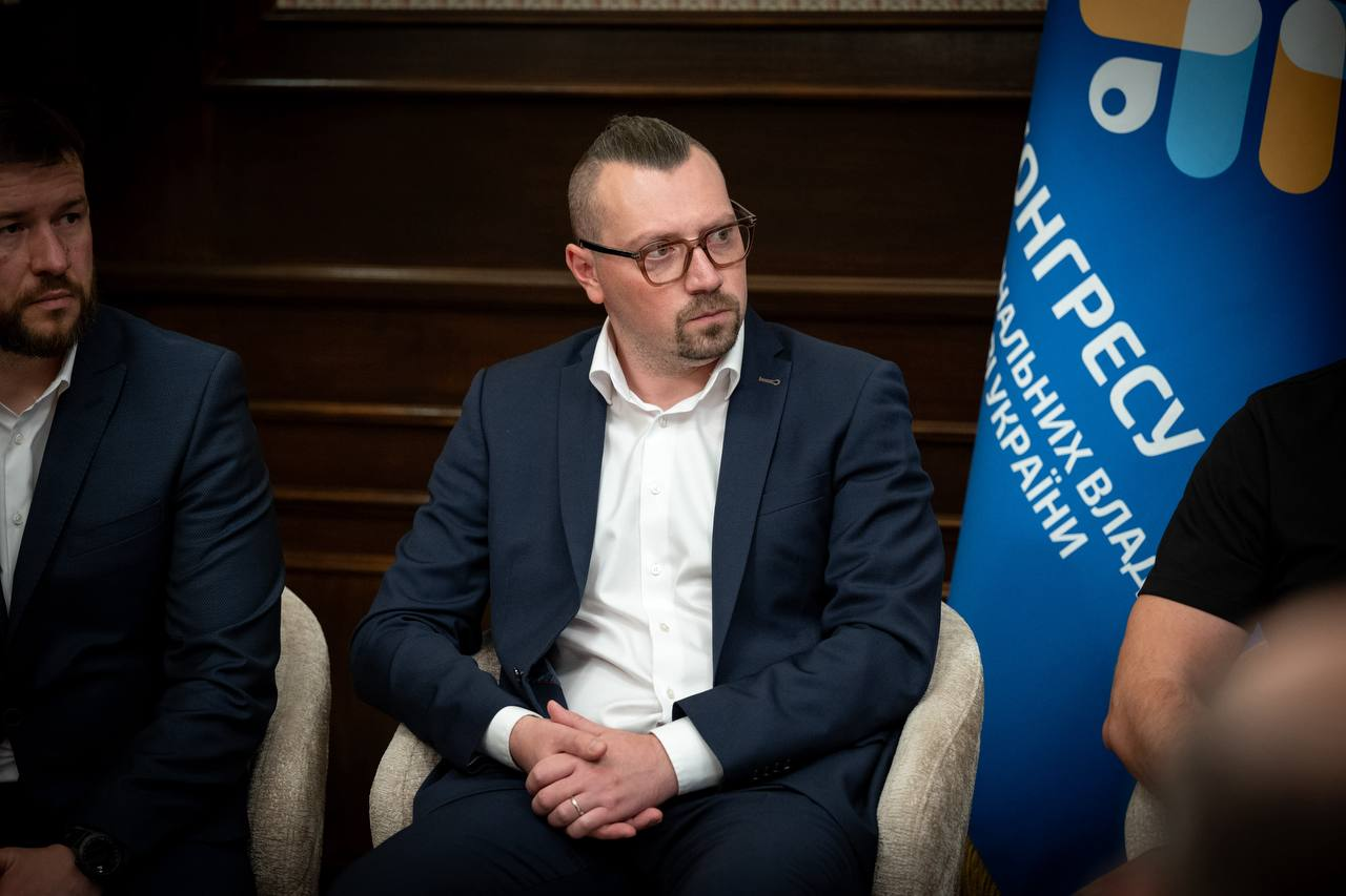 Міністр соціальної політики, сім'ї та єдності України Денис Улютін (ліворуч, у темному костюмі та білій сорочці) сидить у кріслі, повернувшись у профіль, та уважно слухає колег поруч із Міністром у справах громад, територій та внутрішньо переміщених осіб України Віталієм Безгіним (по центру, в окулярах, класичному темно-синьому костюмі та білій сорочці) під час засідання Конгресу місцевих та регіональних влад при Президентові України. Віталій Безгін зафіксований із зчепленими пальцями рук перед собою під час обговорення ситуації з державними фінансами, праворуч на задньому плані встановлено синій стяг Конгресу з круговою різнокольоровою емблемою та чітким білим написом «КОНГРЕСУ місцевих та регіональних влад при Президентові УКРАЇНИ», а в інтер'єрі приміщення видно класичні темні дерев'яні стінові панелі.