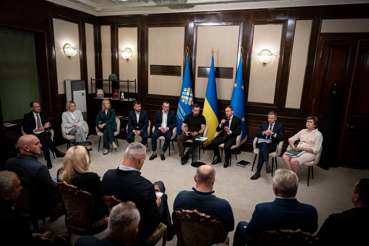 Учасники засідання Конгресу місцевих та регіональних влад при Президентові України розмістилися півколом у залі з класичними дерев'яними стіновими панелями та витонченими настінними світильниками-бра, де у центрі за окремим столом з мікрофоном у руках виступає Прем'єр-міністр України Сергій Корецький, ліворуч від якого сидить заступник керівника Офісу Президента України Віктор Микита у чорній футболці, а далі в ряд у кріслах розмістилися Міністр у справах громад, територій та внутрішньо переміщених осіб України Віталій Безгін (п'ятий ліворуч) та Міністр соціальної політики, сім'ї та єдності України Денис Улютін (четвертий ліворуч). Праворуч від центрального столу сидить Міністр фінансів України Сергій Марченко в картатому темно-синьому костюмі, поруч із яким з правого краю розмістилася учасниця засідання у світлому вбранні з зеленою папкою на колінах, на задньому плані між промовцями встановлено Державний прапор України, синій стяг Конгресу з круговою різнокольоровою емблемою та прапор Європейського Союзу із золотими зірками, а на передньому плані спиною до кадру сидять голови громад та народні депутати, серед яких ліворуч виділяються Миколаївський міський голова Олександр Сєнкевич (крайній ліворуч, чоловік у піджаку) та мер Києва Віталій Кличко (третій ліворуч, чоловік міцної статури).