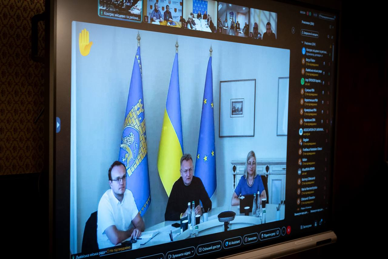 Великий монітор у залі відображає вікно відеотрансляції з кабінету Львівської міської ради під час засідання Конгресу місцевих та регіональних влад при Президентові України, де по центру за круглим столом виступає Львівський міський голова Андрій Садовий у чорному світшоті. Ліворуч від нього сидить перший заступник у білій сорочці-поло та окулярах, а праворуч — учасниця засідання у синій блузці та окулярах біля відкритого ноутбука. Позаду промовців на флагштоках підняті прапори: офіційний синій стяг міста Львова із золотим зображенням лева та міської брами, Державний прапор України та прапор Європейського Союзу.