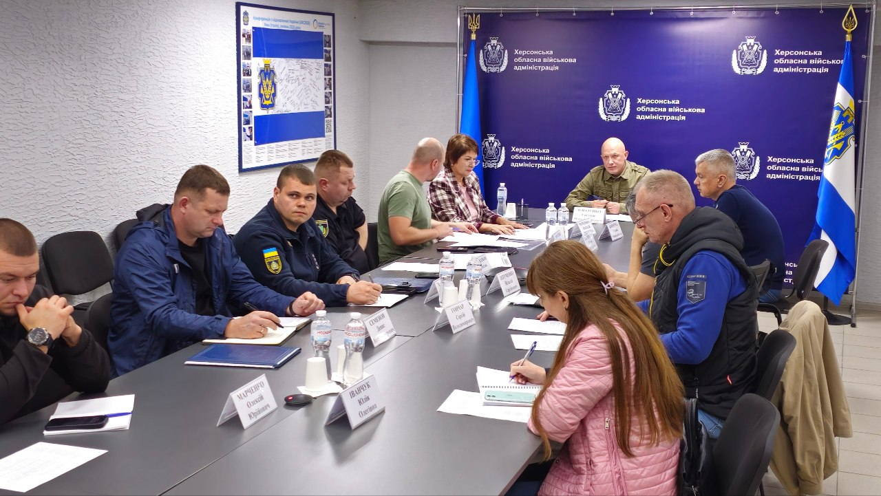 Заступник начальника Херсонської ОВА Євгеній Ігнатенко (у центрі на задньому плані, в камуфляжній формі) проводить засідання обласної комісії з питань евакуації в офіційному кабінеті, де ліворуч від нього за столом сидить заступниця начальника Херсонської МВА Наталія Чорненька у чорно-білій картатій сорочці, а представники військових адміністрацій, залізничники та правоохоронці розмістилися по обидва боки довгого сірого Т-подібного столу й опрацьовують робочі матеріали щодо безпекової ситуації в деокупованих громадах і логістики вивезення людей. У торці залу ліворуч від Євгенія Ігнатенка на флагштоці піднято Державний прапор України, праворуч — прапор Херсонської області, а позаду встановлено великий темно-синій брендований банер із повторюваними білими гербами та написами «Херсонська обласна військова адміністрація». На столі перед кожним учасником розставлено іменні паперові таблички, пляшки з мінеральною водою та розкладено папки з паперами, при цьому ліворуч у ряд сидять чоловік у чорній кофті з годинником на руці, посадовець у синій куртці, військовослужбовець у камуфляжній формі із нашитим синьо-жовтим прапором на плечі та інші учасники, а праворуч на передньому плані директорка Департаменту здоров'я Херсонської ОВА Тетяна Карчевич у рожевій кофті робить записи в блокноті поруч із сивочолим чоловіком в окулярах та синьому жилеті з шевроном ОДА, водночас на світлій бічній стіні ліворуч закріплено великий плакат-інфографіку із зображенням прапора Херсонської області.