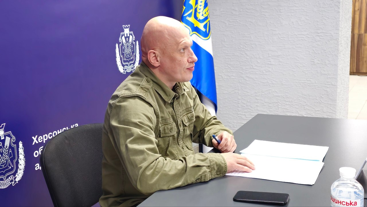 Заступник начальника Херсонської обласної військової адміністрації Євгеній Ігнатенко сидить за сірим робочим столом у кабінеті, тримаючи в правій руці синю кулькову ручку та зосереджено даючи вказівки під час засідання обласної комісії з питань евакуації. Перед посадовцем на столі розкладено паперові бланки, документи та лежить чорний мобільний телефон, у нижньому правому кутку помітно пластикову пляшку мінеральної води, а сам керівник показаний у профіль на тлі великого темно-синього брендованого банера з білими емблемами-гербами та частковим написом «Херсонська обласна державн адміністрація», праворуч від якого встановлено прапор Херсонської області під світло-сірою текстурною стіною.