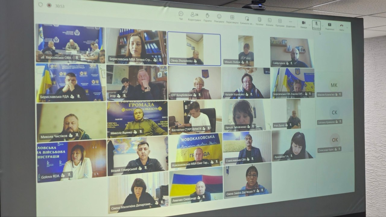 Великий прямокутний проекційний екран у залі засідань відображає вікно онлайн-конференції під час проведення засідання обласної комісії з питань евакуації під головуванням заступника начальника Херсонської ОВА Євгенія Ігнатенка, до якого долучились керівники профільних структурних підрозділів ХОДА, правоохоронці, представники та начальники ХМВА, районних та військових адміністрацій населених пунктів області, а також залізничники. Вони показані у віконцях відеотрансляцій із робочих кабінетів на тлі Державних прапорів України та брендованих стягів, опрацьовуючи питання логістики та безпеки пунктів відправлення людей.