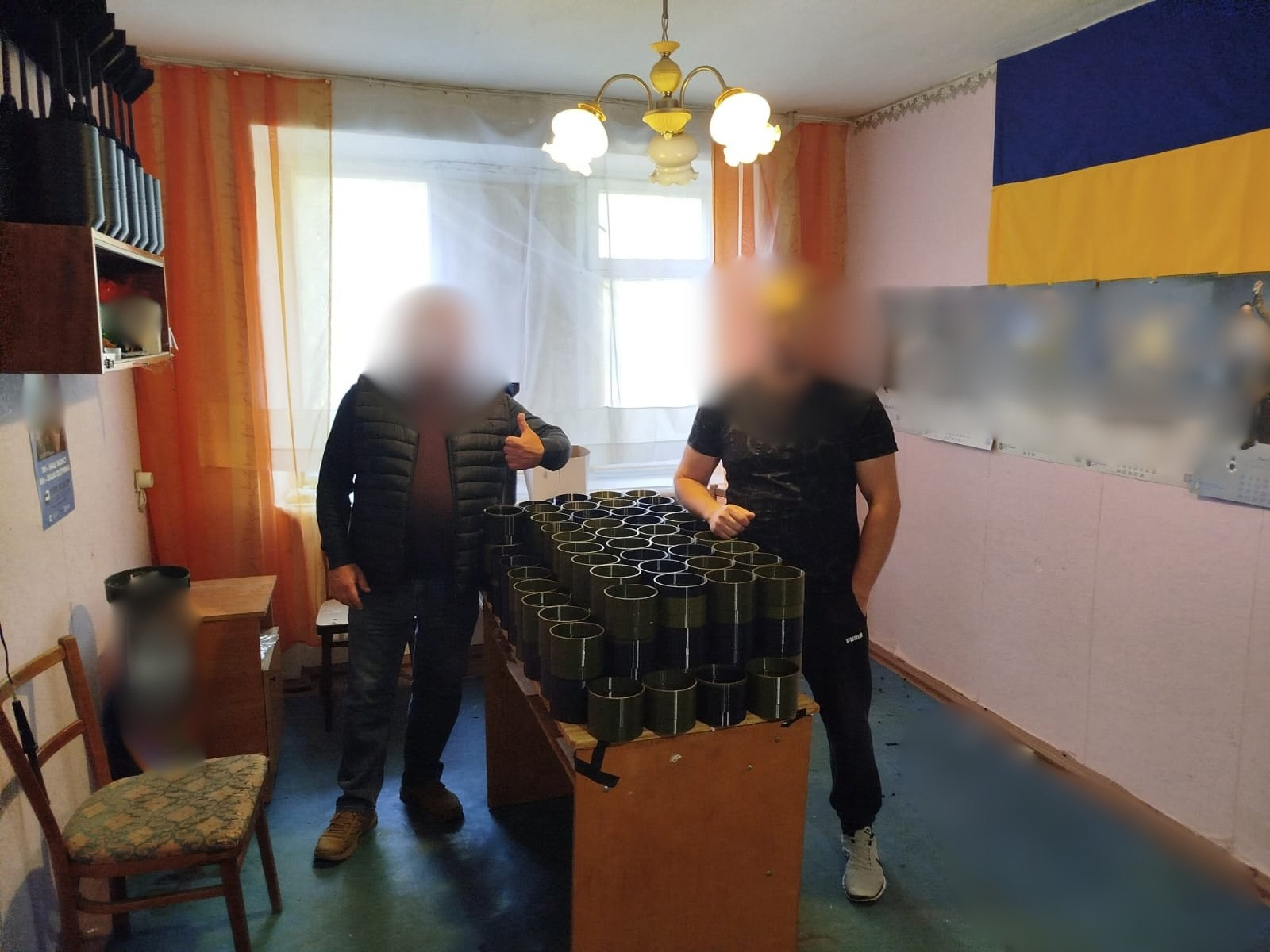 Двоє чоловіків із розмитими з міркувань безпеки обличчями стоять у світлому робочому кабінеті по обидва боки дерев'яного столу, на якому рівними рядами виставлено велику кількість виготовлених на 3D-принтері військових виробів захисного зеленого кольору, що стало результатом об'єднання зусиль Виноградівської та Чулаківської громад задля підтримки Сил оборони України. Чоловік ліворуч одягнений у темну балонову жилетку, сині джинси та коричневе взуття, він піднімає вгору великий палець лівої руки на знак успішного завершення збору, а чоловік праворуч у чорній футболці та темних спортивних штанах спирається правою рукою на край столу з готовою продукцією, для закупівлі компонентів та пластику якої працівники Виноградівської сільської військової адміністрації провели збір коштів, а фахівці Чулаківської СВА реалізували сам процес друку. На задньому плані кадру велике світле вікно завішене прозорим тюлем та помаранчевими шторами, під стелею горить люстра, на рожевій стіні праворуч закріплено великий синьо-жовтий Державний прапор України та інформаційні стенди, які розмито, ліворуч у кабінеті розміщено стілець із м'якою оббивкою, тумбу та відкриті настінні полиці.
