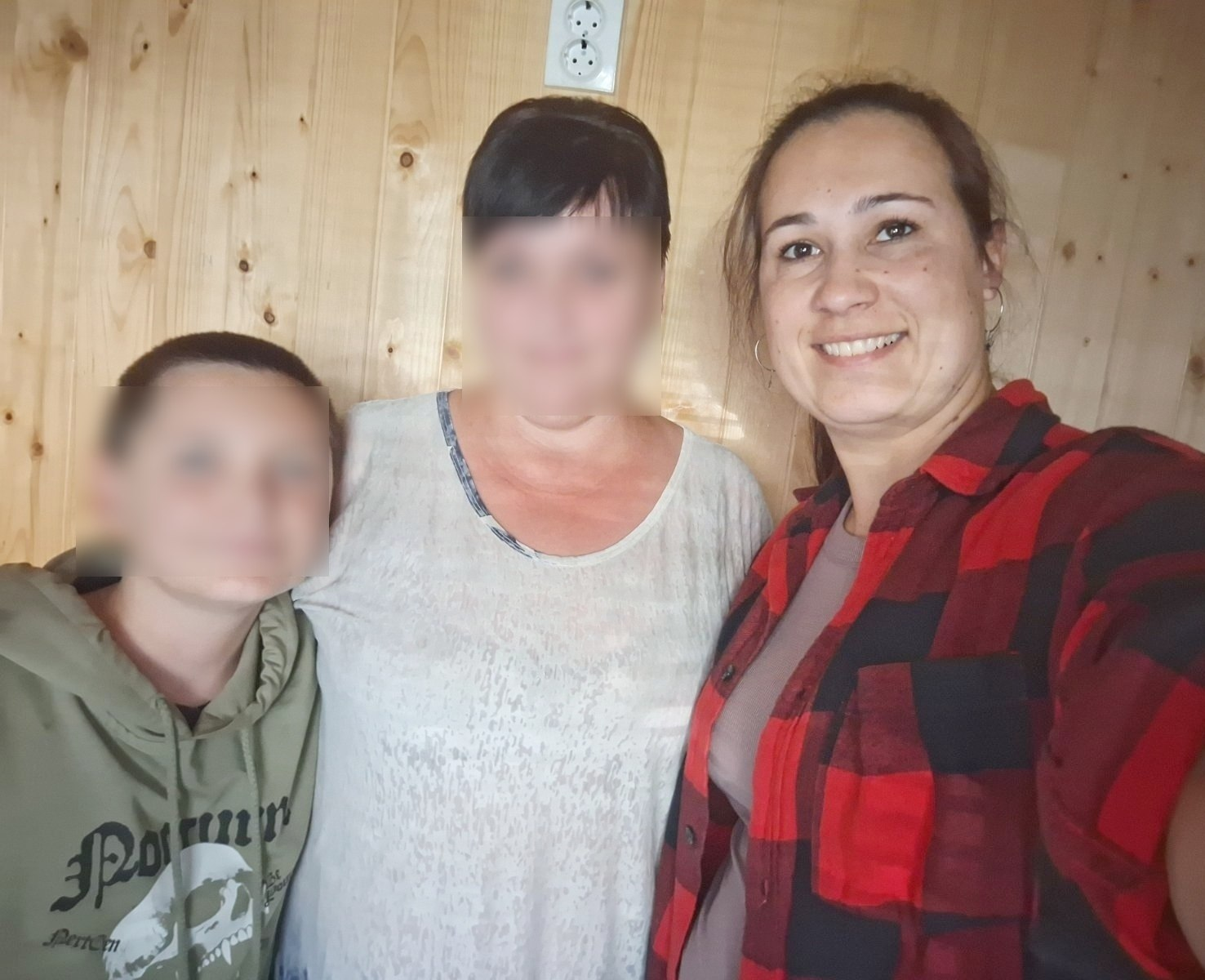 Начальниця служби у справах дітей Таврійської міської ради (праворуч, з усмішкою, у червоно-чорній картатій сорочці) робить спільне селфі-фото під час відвідин родини з дитиною, яку разом із мамою евакуювали з тимчасово окупованих територій Херсонщини до Київської області. У центрі кадру стоїть мати хлопчика у світлій футболці з дрібним візерунком на комірі, а ліворуч до неї тулиться її син у хакі-зеленому худі з написами й малюнком на грудях, причому обличчя дитини та жінки розмиті з міркувань безпеки, а сама зустріч присвячена супроводу родини, перевірці умов проживання та наданню сприяння у розв’язанні актуальних питань після тривалого перебування в окупації. Усі троє зафіксовані крупним планом у кімнаті на тлі стіни з вертикальних світло-коричневих дерев'яних дощок.