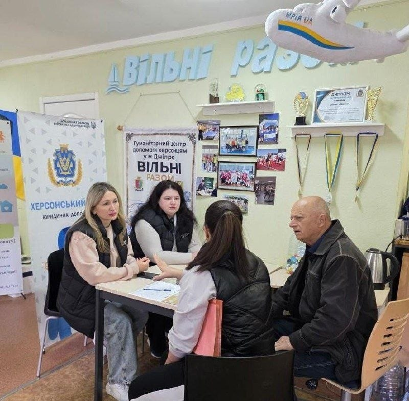 Чотири людини — дві співробітниці-консультантки (сидять по ліву сторону столу) та двоє відвідувачів (сидять по праву сторону) — зображені за робочим столом у світлому кабінеті гуманітарного центру «Вільні разом» у Дніпрі під час надання юридичних консультацій та послуг соціального супроводу для ВПО з Херсонщини. Ліворуч за столом сидить перша консультантка у світлому худі та чорній жилетці, яка веде бесіду, а поруч із нею праворуч сидить її колега у світлій кофті та чорному жилеті, навпроти них по ліву сторону столу розмістилися жінка спиною до кадру з рожевою папкою в руках та праворуч літній чоловік у темній куртці, які уважно слухають роз'яснення фахівців. На задньому плані світло-зелена стіна прикрашена великим блакитним написом «ВІЛЬНІ РАЗОМ», плакатом, рамками з фотографіями, кубками на полицях та м'якою іграшкою у формі літака «МРІЯ UA», тоді як ліворуч у кімнаті встановлено білий банер «ХЕРСОНСЬКИЙ ХАБ ЮРИДИЧНА ДОПОМОГА».