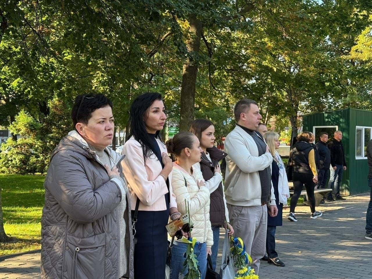 Група представників координаційного центру «Вільні разом» у Дніпрі стоїть у ряд під час урочистого молебню та покладання квітів на Алеї Слави, притиснувши праві руки до грудей під час виконання Державного Гімну України до Дня захисників і захисниць України. На передньому плані ліворуч стоїть жінка в сірій куртці, поруч із нею — друга учасниця заходу в ніжно-рожевому піджаку та чорній спідниці, далі по центру розмістилася третя жінка у світлій куртці в ромбик, яка тримає в руках невеликий пакуночок, а за нею стоїть четверта дівчина у коричневому жилеті поверх білої кофти. Чоловік у білому кардигані, сірих штанах та чорній футболці стоїть праворуч від них, тримаючи внизу букет синьо-жовтих квітів, а поруч у ряд вишикувалися інші учасники та відвідувачі, водночас на задньому плані кадру на тлі великих зелених дерев паркової зони видно перехожих, лавки та невелику зелену господарську будівлю.