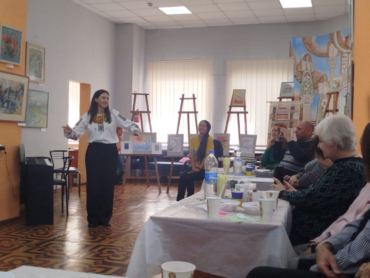 Жінка у красивій білій вишиванці з традиційним орнаментом та довгій чорній спідниці емоційно виступає, розвівши руки в сторони, під час творчої зустрічі «Майстер сонячного пензля» для переселенців з Херсонщини в Одесі, яка присвячена творчості художниці Лариси Дем’янишиної. Поруч у залі на стільці сидить інша дівчина у жовтій футболці та уважно слухає промову, водночас праворуч за довгим столом із частуваннями та паперовими стаканчиками розмістилися глядачі похилого віку й діти, які спостерігають за виступом та фотографують захід. Зустріч для ВПО з Верхньорогачицької громади проходить у світлому мистецькому залі з паркетною підлогою, де на задньому плані вздовж вікон на дерев'яних мольбертах виставлено картини й художні малюнки, на стіні ліворуч розвішані репродукції в рамках, а праворуч облаштовано велику тематичну виставку літератури та ілюстрацій.