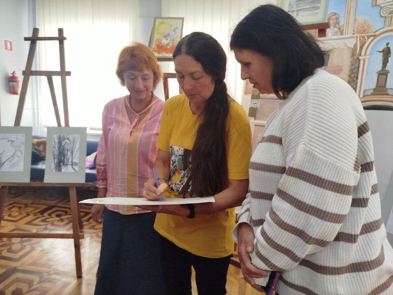 Художниця Лариса Дем’янишина в яскравій жовтій футболці з принтом та з довгим темним волоссям стоїть по центру мистецького залу і зосереджено робить авторський підпис або малює ручкою на великому білому аркуші паперу, який тримає в руках, під час творчої зустрічі «Майстер сонячного пензля» для переселенців із Херсонщини в Одесі. З лівого боку від неї за процесом з усмішкою спостерігає жінка в рожево-жовтій смугастій кофті та довгій темній спідниці, а праворуч нахилилася інша учасниця заходу в білому светрі в широку коричневу смужку. Спільна творча взаємодія ВПО з Верхньорогачицької громади та мисткині відбувається у світлому приміщенні бібліотеки з візерунчастим дерев'яним паркетом на тлі виставкових дерев'яних мольбертів із малюнками, настінних художніх робіт, декоративного банера із зображенням Пам'ятника Дюку де Рішельє та інших елементів мистецького оформлення кімнати.