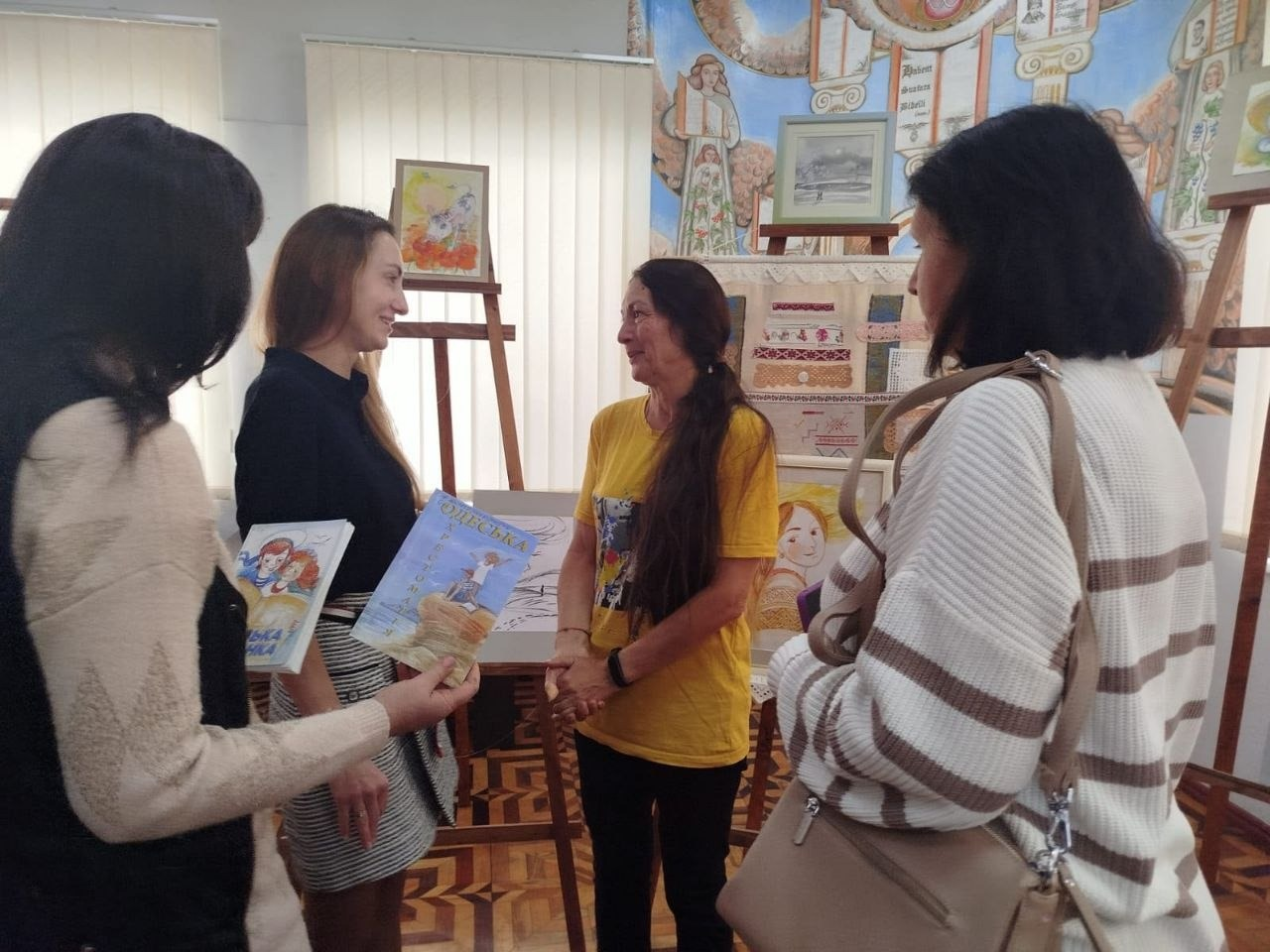 Художниця Лариса Дем’янишина в яскравій жовтій футболці з принтом та з довгим темним волоссям стоїть по центру мистецького залу і тепло спілкується з відвідувачками творчої зустрічі «Майстер сонячного пензля» для переселенців із Херсонщини в Одесі. Перед нею стоїть дівчина у чорній кофті та світлій спідниці в дрібну смужку, яка тримає в руках дитячі книги одеського видавництва, зокрема видання під назвою «Одеська», ліворуч спиною до кадру зафіксована учасниця в бежевому светрі, а праворуч наближається жінка у білому светрі в широку коричневу смужку з бежевою сумкою на плечі. Зустріч ВПО з Верхньорогачицької громади з мисткинею проходить у світлому приміщенні бібліотеки з візерунчастим дерев'яним паркетом на тлі виставкових дерев'яних мольбертів із малюнками, настінних художніх робіт та великого декоративного панно з етнічними мотивами.