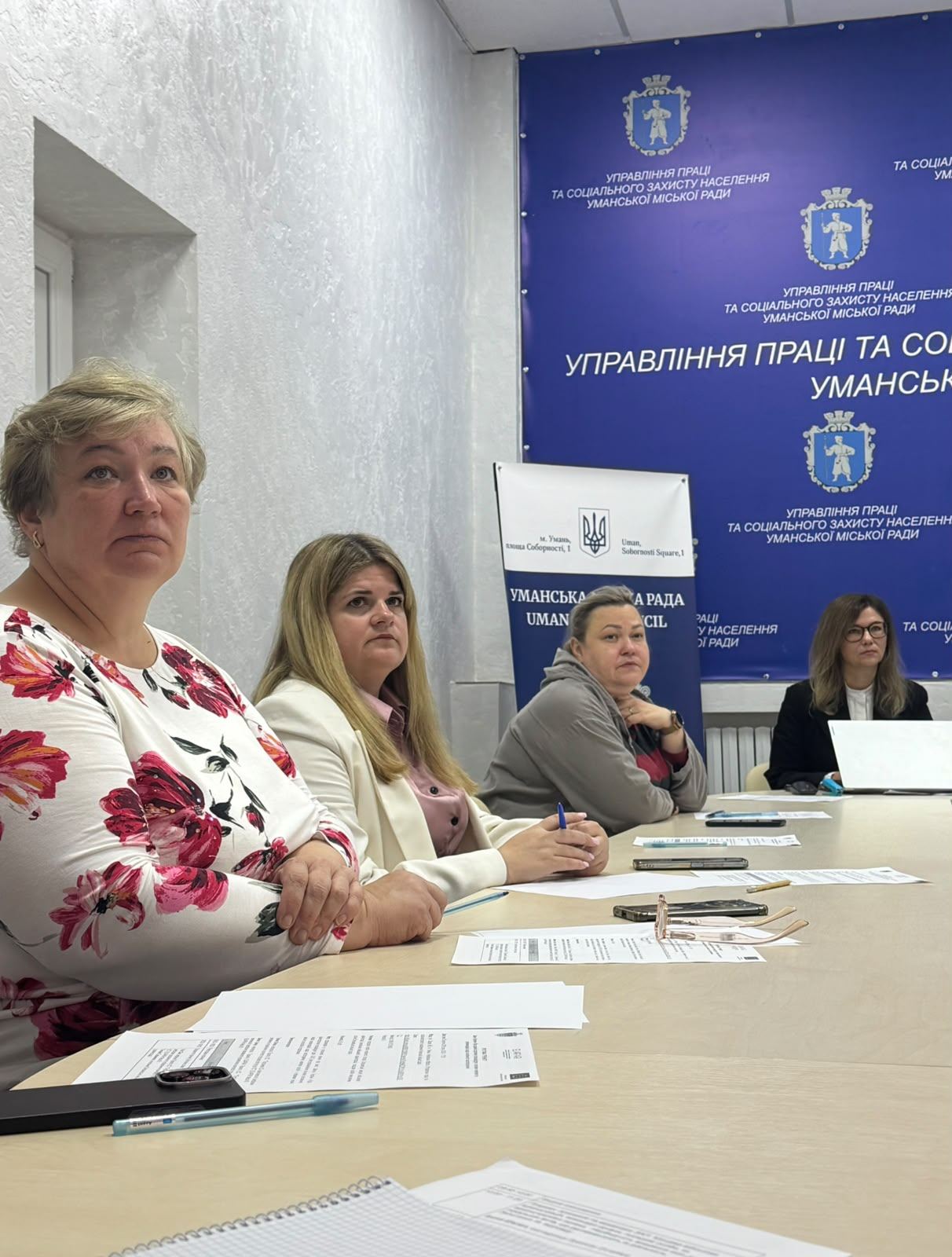 Чотири учасниці тренінгу щодо застосування Закону України «Про адміністративну процедуру» в межах проєкту Європейського Союзу (EU4PAR III) сидять у ряд за світло-бежевим робочим столом в Управлінні праці та соціального захисту населення Уманської міської ради й уважно слухають виступи колег. На передньому плані ліворуч сидить жінка в білій блузці з великим яскраво-рожевим квітковим принтом, поруч із нею — посадовиця у світло-бежевому жакеті та рожевій блузці, далі по центру — учасниця у сірій спортивній кофті з капюшоном, яка тримає синю ручку, а праворуч на задньому плані сидить інша фахівчиня в окулярах та темному одязі біля відкритого білого ноутбука. Перед присутніми на столі розкладено робочі документи, бланки для нотаток та мобільний телефон, водночас на задньому плані встановлено великий темно-синій брендований банер із білими гербами міста та частковим білим написом «УПРАВЛІННЯ ПРАЦІ ТА СОЦІАЛЬНОГО ЗАХИСТУ...», ліворуч від якого видно білу інформаційну стійку Уманської міської ради.