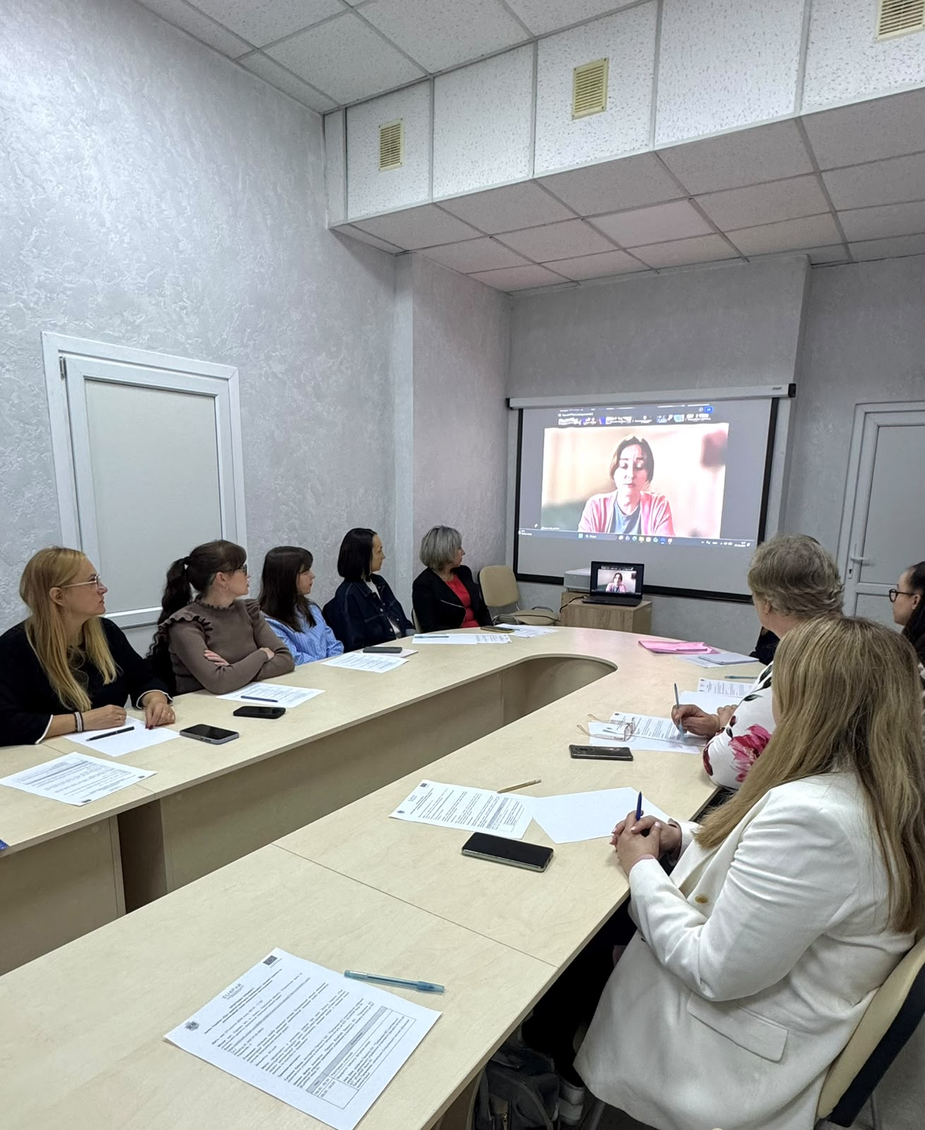 Великий прямокутний проекційний екран відображає вікно онлайн-конференції Zoom з виступом експертки-юристки під час тренінгу щодо застосування Закону України «Про адміністративну процедуру» в межах проєкту Європейського Союзу (EU4PAR III). Фахівці Олешківської МВА та міської ради сидять довкола великого світло-бежевого столу й уважно слухають лекцію. По ліву сторону в ряд розмістилися учасниці, серед яких дівчата в коричневому светрі, блакитній сорочці та чорній куртці, а по праву сторону столу сидить решта колективу, зокрема на передньому плані видно посадовицю у світлому жакеті та жінку в блузці з великим рожевим квітковим принтом. На столі перед присутніми розкладено бланки документів із логотипом проєкту ЄС, чисті аркуші для нотаток, ручки та телефони. Зустріч проходить у світлому кабінеті з підвісною стелею та білими дверима з боків залу.