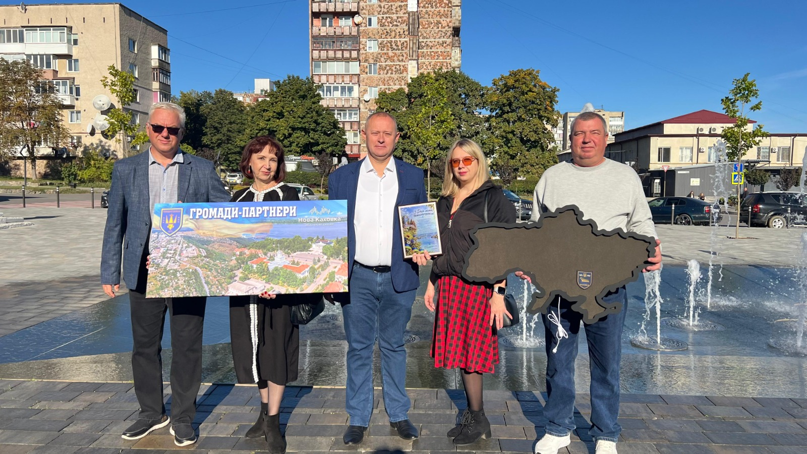 Керівництво Новокаховської МВА та Новодністровської міської ради спільно тримають пам'ятні банери та символічну дерев'яну мапу України під час робочого візиту начальника Новокаховської міської військової адміністрації Олега Тарабаки до Новодністровська у межах національного проєкту «Пліч-о-пліч: згуртовані громади». Учасники делегації стоять на міській площі біля фонтана, що працює, де по центру розмістився начальник Новокаховської МВА Олег Тарабака у білій сорочці та синьому піджаку, ліворуч від якого жінка тримає великий кольоровий плакат із гербом Нової Каховки та написом «ГРОМАДИ-ПАРТНЕРИ Нова Каховка», а крайнім ліворуч стоїть чоловік в окулярах та синьому класичному піджаку. Праворуч від Олега Тарабаки перебуває міська голова Новодністровська Наталя Цимбалюк у червоно-чорній картатій спідниці та сонцезахисних окулярах із пам'ятною книгою в руках, а крайнім праворуч розмістився чоловік у світлій кофті, який утримує об'ємний темний макет карти України із гербом. На задньому плані кадру під безхмарним синім небом височіє багатоповерховий житловий будинок коричневого кольору, ростуть зелені дерева, облаштовано пішохідну зону з бруківки та видно припарковані автомобілі біля міських будівень.