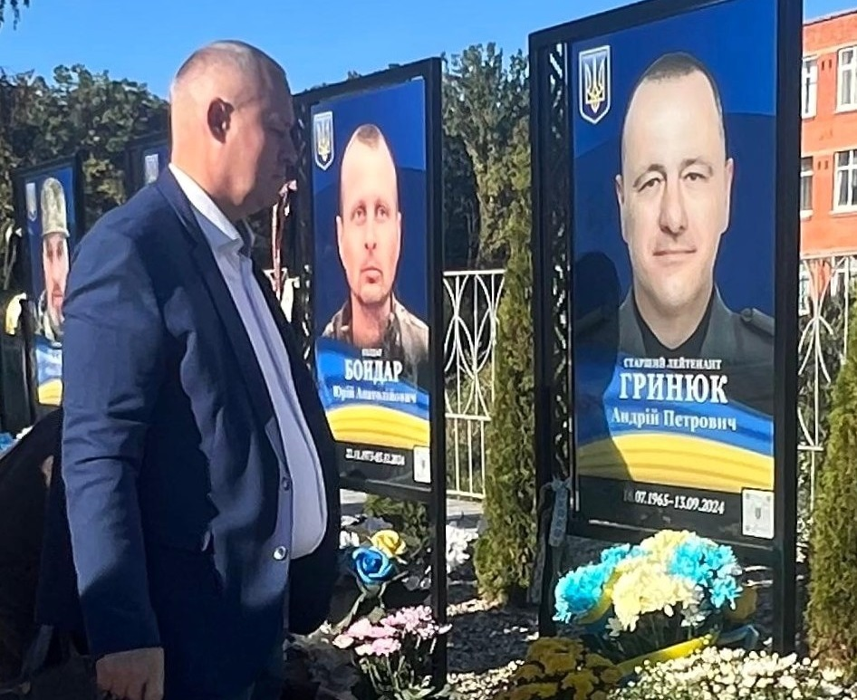 Начальник Новокаховської МВА Олег Тарабака в класичному синьому піджаку та білій сорочці стоїть у профіль, схиляючи голову під час хвилини мовчання на Алеї Слави в Новодністровську біля меморіального стенда з портретом полеглого захисника України, старшого лейтенанта Андрія Петровича Гринюка. Праворуч на великому чорному металевому стенді розміщено велику фотографію новокаховчанина Андрія Гринюка у військовій формі з Державним Гербом та синьо-жовтим прапором у нижній частині, під якими встановлено кошик із блакитними та жовтими квітами, а ліворуч на задньому плані видно аналогічний пам'ятний стенд з портретом іншого полеглого воїна Юрія Анатолійовича Бондаря та обриси цегляної будівлі за металевим парканом на тлі зелених дерев.