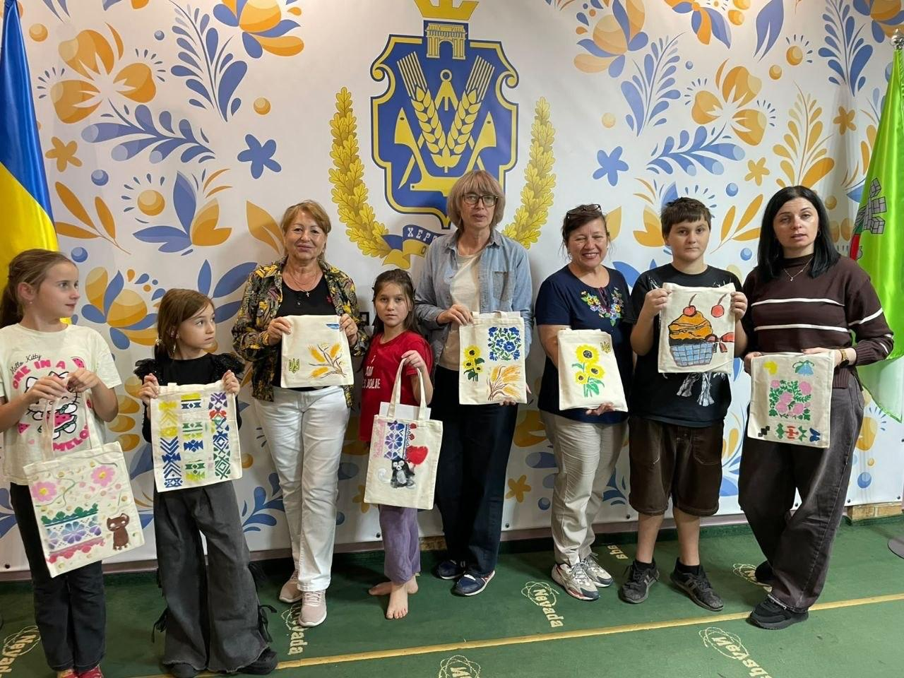 Група дітей та дорослих із числа ВПО з Херсонщини позують для спільного пам'ятного фото, демонструючи власноруч розписані тканинні екосумки-шопери після завершення творчого майстер-класу в координаційному центрі «Вільні разом» в Ужгороді. Учасники вишикувалися в один ряд на тлі великого білого банера, який щедро прикрашений яскравими жовто-блакитними рослинними орнаментами в народному стилі та великим офіційним гербом Херсонської області по центру. З лівого боку стоять троє дівчаток у світлих та червоній футболках, поруч із якими позують три дорослі жінки-учасниці, тримаючи екосумки з малюнками соняшників, квітів та традиційних візерунків, а праворуч розмістилися хлопчик у чорній футболці з малюнком кексу на шопері та майстриня-організаторка в коричневому смугастому светрі з темним волоссям.