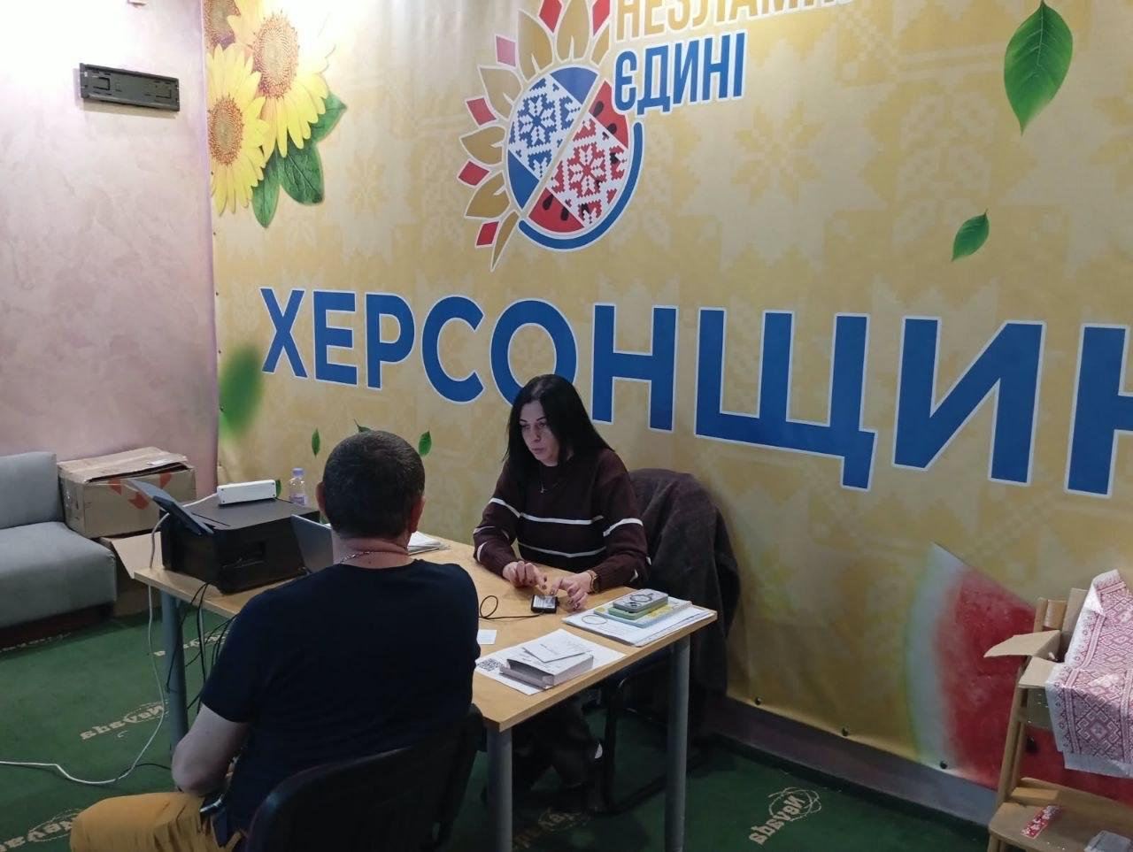 Співробітниця-консультантка з темним волоссям у коричневому смугастому светрі сидить за робочим столом та надає правову консультацію чоловікові-переселенцю в чорній футболці у координаційному центрі «Вільні разом» в Ужгороді. На столі розкладено паперові документи, брошури та телефони, ліворуч встановлено чорний принтер, а сама індивідуальна зустріч від представника Міжнародного фонду «Регіон Карпат» (NEEKA) проходить на тлі великого жовтого брендованого банера з малюнками соняшників, кавуна та великими написами «ХЕРСОНЩИНА», «ЄДИНІ» та «НЕЗЛАМНА». Робоче місце облаштоване в кімнаті з зеленим ковроліном, де ліворуч помітно край сірого дивана, а праворуч — дитячий стільчик із вишитим рушником.