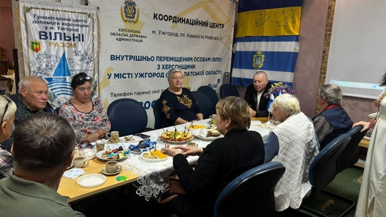Велика група внутрішньо переміщених осіб похилого віку з Херсонщини сидить довкола святкового столу з фруктами та солодощами під час вечорниць «Вечір зустрічі у теплому колі», організованих до Міжнародного дня людей старшого віку в координаційному центрі «Вільні разом» в Ужгороді. Літні мешканці затишно спілкуються за чаєм на тлі офіційних банерів гуманітарного хабу та Херсонської обласної державної адміністрації з гербом, а у правому кутку кімнати підвішено підписаний Державний прапор України та синьо-білий стяг із гербом міста Херсона.