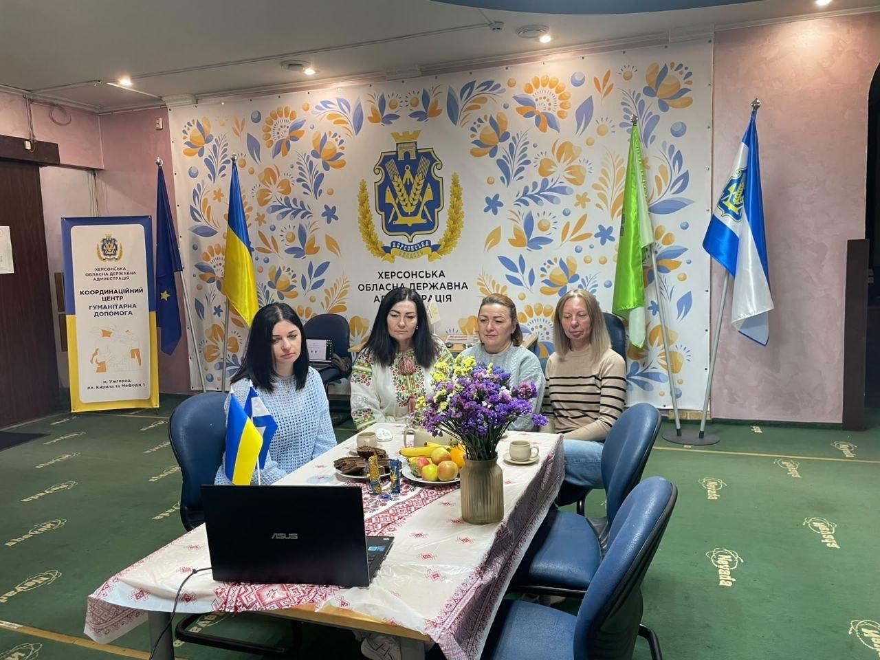 Чотири співробітниці координаційного центру «Вільні разом» в Ужгороді сидять за столом перед відкритим ноутбуком та беруть участь в онлайн-конференції з нагоди ХХ ювілейного Міжнародного фестивалю театрів ляльок «Інтерлялька». На столі, який застелено скатертиною з червоним народним орнаментом, виставлено вазу з букетом фіолетових квітів, тарілку з фруктами, печиво та чашки, зліва направо сидять: дівчина у блакитній кофті з маленьким прапорцем України, далі в ряд розмістилися її колеги у вишиванці, блакитному светрі та коричневій кофті в смужку. Зустріч проходить у залі з зеленим ковроліном на тлі великого білого банера з патріотичним орнаментом, гербом та написом «ХЕРСОНСЬКА ОБЛАСНА ДЕРЖАВНА АДМІНІСТРАЦІЯ», водночас з лівого краю піднято Державний прапор України й встановлено жовтий ролап-стенд, а з правого боку піднято прапор міста Ужгорода та прапор Херсонської області.