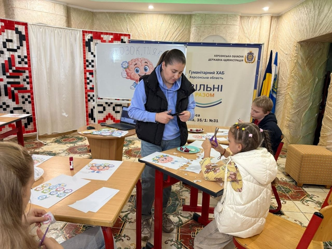 Жінка-організаторка в чорній жилетці поверх блакитного худі стоїть по центру дитячої кімнати одеського координаційного центру «Вільні разом» і допомагає дітям вирізати деталі для аплікацій ножицями під час творчого майстер-класу «Мозок, ти як?» у межах Всеукраїнської програми ментального здоров’я. Навколо парт у світлому залі з візерунчастою плиткою на підлозі розмістилися юні переселенці з Херсонщини, серед яких на передньому плані дівчинка в білій жилетці акуратно працює ножицями, праворуч за столом сидить хлопчик у чорному світшоті, а ліворуч дівчинка вирізає квіти-емоції. На столах розкладено чисті аркуші паперу, клеєві олівці, шаблони та зразки малюнків, водночас на задньому плані встановлено білу магнітну дошку з плакатом «Мозок, ти як?», великий офіційний банер Гуманітарного хабу Херсонської області «Вільні разом», білі ширми з традиційним червоно-чорним вишитим орнаментом, а у правому кутку кімнати на флагштоках піднято Державний прапор України та прапор Херсонської області.
