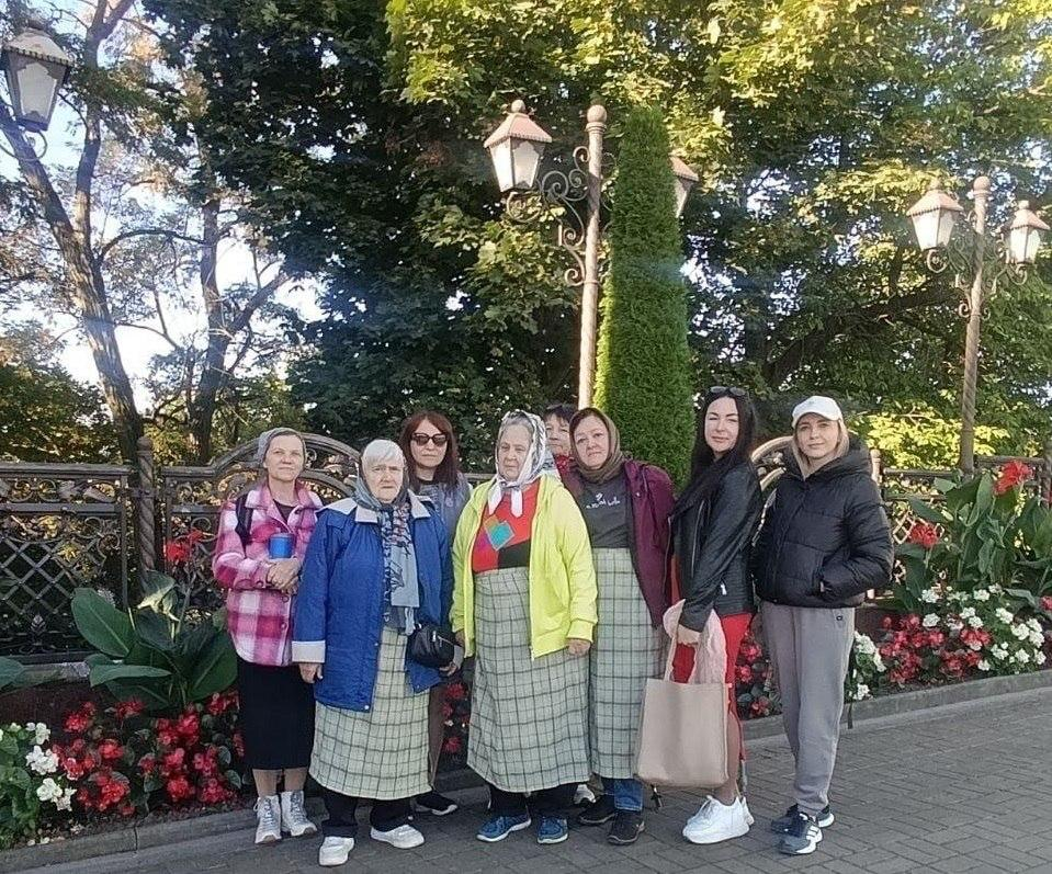 Група переселенок із Херсонщини різного віку позує для спільного пам'ятного фото на вимощеній бруківкою доріжці під час екскурсії до села Банчени та відвідин монастиря, організованих координаційним центром «Вільні разом» у Чернівцях. Жінки вишикувалися в ряд на тлі металевого декоративного паркану та високих зелених дерев, де ліворуч стоїть учасниця в рожево-білій картатій сорочці, поруч із нею — літня жінка у синій куртці та світлій довгій спідниці у клітинку, далі по центру розмістилися відвідувачки в сонцезахисних окулярах, у яскравій жовтій кофті та бордовій куртці, а праворуч стоять дві молодші жінки у чорній шкіряній куртці з бежевою сумкою та у чорному пуховику зі світлою кепкою на голові. На задньому плані вздовж паркану встановлено старовинні ліхтарі на високих металевих стовпах, росте струнка зелена туя, а біля ніг екскурсанток облаштовано красиві клумби з квітучими червоними й білими бегоніями.