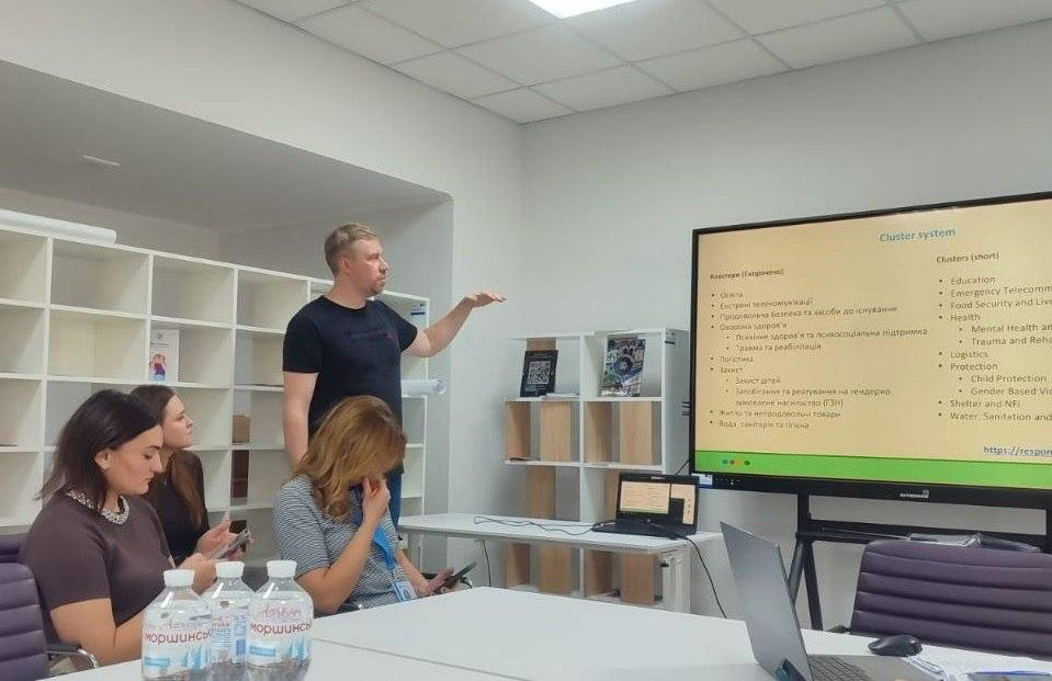 Чоловік-спікер у чорній футболці стоїть біля великого інтерактивного екрана й активно жестикулює правою рукою, презентуючи інформацію про кластерну систему «Cluster system» для фахівчинь координаційного центру «Вільні разом» у Чернівцях під час зустрічі волонтерських організацій у Департаменті соціального захисту населення Чернівецької ОВА. На великому дисплеї виведено слайд із переліком напрямів роботи українською та англійською мовами, зокрема «Освіта», «Екстренні телекомунікації», «Охорона здоров'я» та «Психічне здоров'я та психосоціальна підтримка». Перед екраном на білому столі розташований відкритий чорний ноутбук, а ліворуч за основним столом сидять учасниці заходу із херсонського хабу, які уважно слухають доповідача та роблять нотатки в телефонах, при цьому на передньому плані виставлено пляшки мінеральної води «Моршинська». Зустріч проходить у світлому сучасному кабінеті з підвісною стелею, великим білим стелажем із полицями для документів на задньому плані та м'якими офіційними кріслами фіолетового кольору.