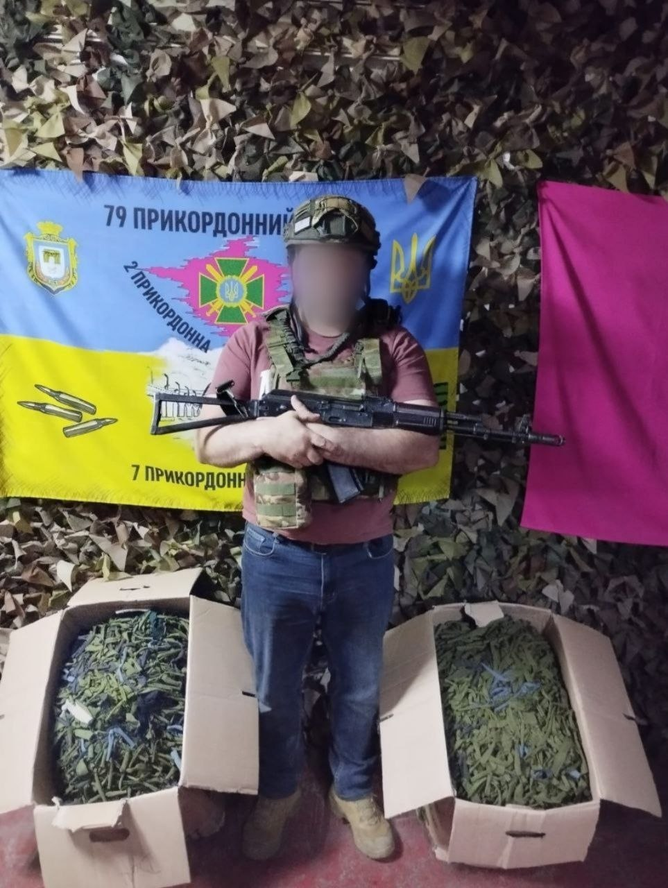 Український військовослужбовець у камуфляжному шоломі, захисному бронежилеті розвантаження та синіх джинсах стоїть на повний зріст і тримає в руках автомат, приймаючи чергову допомогу від Чаплинської та Долматівської громад для Сил оборони України. Обличчя бійця розмите з міркувань безпеки, а сам він перебуває у приміщенні на тлі великого синьо-жовтого прапора 79 прикордонного загону Державної прикордонної служби України з написами «79 ПРИКОРДОННИЙ», «2 ПРИКОРДОННА» та «7 ПРИКОРДОННИЙ», який закріплений на стіні під маскувальною сіткою поруч із рожевим полотном. Біля ніг захисника на підлозі виставлено дві великі картонні коробки, які вщент заповнені виготовленими маскувальними халатами зеленого та захисного кольорів, що були передані у підрозділ для виконання завдань на Херсонському та Донецькому напрямках.