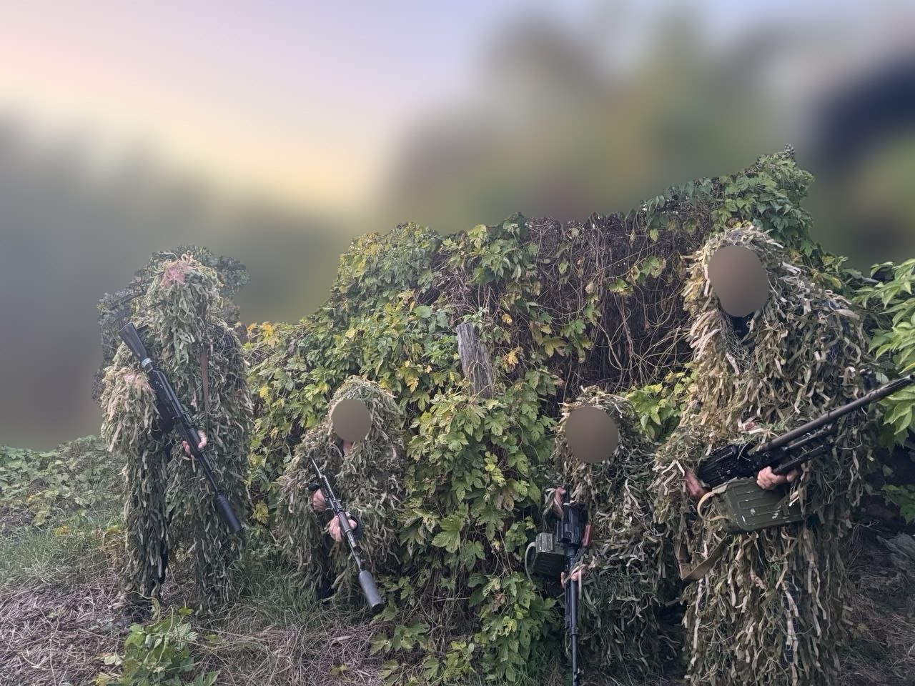 Українські військовослужбовці в одягнених маскувальних халатах («кікіморах») та зі зброєю в руках позиgeneric на позиціях на тлі зелених чагарників і кущів, демонструючи засоби маскування, виготовлені завдяки співпраці Чаплинської та Долматівської громад із волонтерами. Обличчя бійців розмиті з міркувань безпеки, а самі вони стоять у ряд, повністю зливаючись із навколишньою рослинністю та деревами під вечірнім небом. Військові тримають автомати зі встановленими глушниками та кулемет із патронною коробкою, демонструючи високу якість плетіння захисних костюмів, які ефективно розмивають силует людини на місцевості та надсилаються у підрозділи Сил оборони на Херсонський і Донецький напрямки.