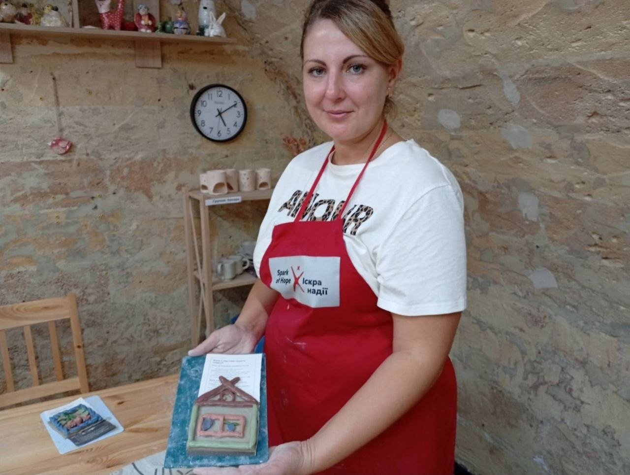 Учасниця майстер-класу в білій футболці з написом та червоному фірмовому фартуху тримає перед собою на блакитній підставці власноруч виліплений із глини декоративний будиночок під час творчого заходу від МБФ «Іскра Надії» та координаційного центру «Вільні разом». Жінка з підібраним волоссям дивиться в кадр, демонструючи готову глиняну плитку у формі хатинки із коричневим дахом, зеленими стінами та двома віконцями. Творче заняття для переселенців Верхньорогачицької громади проходить у кімнаті з автентичними кам'яними стінами, де на задньому плані ліворуч висить круглий настінний годинник, встановлено дерев'яний стелаж із виробами, облаштовано верхню полицю з дрібними сувенірами, а внизу помітно край дерев'яного столу з іншою готовою роботою та стілець.