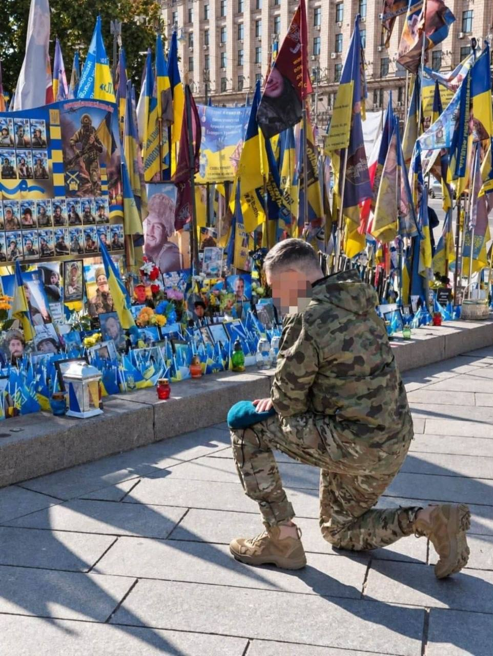 Український військовослужбовець у камуфляжній формі та бежевих тактичних черевиках стоїть на одному коліні, схиливши голову на знак глибокої пошани та скорботи перед народним меморіалом полеглим захисникам України на Майдані Незалежності в Києві під час пам'ятної зустрічі «Герої серед нас». Боєць тримає свій синьо-зелений берет у руці, схилившись на брукованій площі перед гранітним парапетом, який ущент заставлений численними портретами загиблих героїв, лампадками, живими квітами та сотнями маленьких синьо-жовтих прапорців. Позаду меморіальних стендів під яскравим сонцем височіє цілий ліс великих Державних прапорів України та військових стягів із написами, а на дальньому плані височіє монументальна архітектурна будівля в центрі столиці, де переселенці з Херсонщини спільно з координаційним центром «Вільні разом» долучилися до вшанування пам'яті тих, хто віддав життя за свободу Батьківщини.