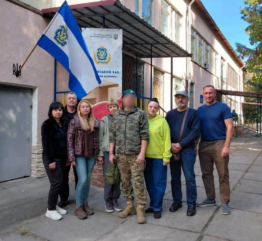 Група цивільних мешканців разом із військовослужбовцем у камуфляжній формі та береті стоять для спільного фото біля входу до координаційного центру «Вільні разом» у Києві під час патріотичної зустрічі «Герої серед нас» до Дня Захисників і Захисниць України. Учасники заходу вишикувалися в один ряд на плитах пішохідної доріжки перед будівлею хабу для ВПО з Херсонщини. З лівого боку від військового стоять три жінки та один чоловік у темній кофті на задньому плані, по центру перебуває захисник із розмитим обличчям, а праворуч від нього розмістилися дівчина у салатовому худі та двоє чоловіків, один із яких в окулярах та кепці із сумкою через плече, а крайній праворуч — у синій футболці. Безпосередньо за присутніми біля входу під навісом встановлено великий білий вертикальний банер із написом «ХЕРСОНСЬКИЙ ХАБ», гербами та зображенням скибки кавуна, ліворуч від якого на стіні будівлі прикріплено малий тризуб та на флагштоці піднято синьо-білий прапор Херсонської області.