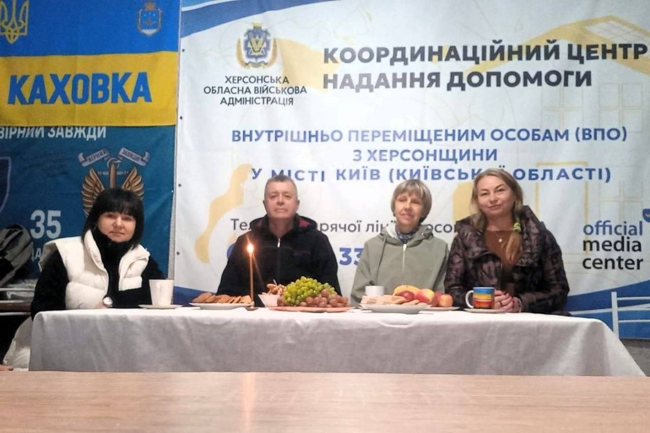 Чотири учасники зустрічі «Герої серед нас» до Дня Захисників і Захисниць України сидять поруч за довгим столом із частуваннями під час надання соціально-психологічної підтримки родинам оборонців у координаційному центрі «Вільні разом» у Києві. На столі, який застелено білою скатертиною, розставлено тарілки з виноградом, печивом, фруктовою нарізкою, чашки для чаю та горить тонка свічка пам'яті, при цьому ліворуч сидить жінка у чорній кофті та білій безрукавці, а далі в ряд розмістилися чоловік у темній куртці, літня жінка у світлій спортивній кофті та жінка у коричневій куртці. Зустріч для внутрішньо переміщених осіб із Херсонщини проходить у світлому кабінеті на тлі великого білого банера із написом «КООРДИНАЦІЙНИЙ ЦЕНТР НАДАННЯ ДОПОМОГИ ВНУТРІШНЬО ПЕРЕМІЩЕНИМ ОСОБАМ (ВПО) З ХЕРСОНЩИНИ У МІСТІ КИЇВ (КИЇВСЬКІЙ ОБЛАСТІ)», ліворуч від якого встановлено жовто-синій прапор Каховки з гербом, написом «КАХОВКА» та гаслом «ВІРНИЙ ЗАВЖДИ».