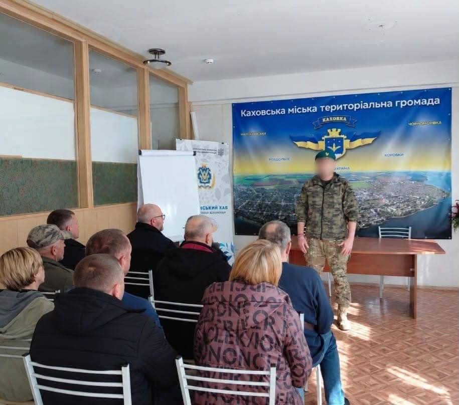 Український військовослужбовець у камуфляжній формі та зеленому береті виступає перед присутніми у залі під час патріотичної зустрічі «Герої серед нас» до Дня Захисників і Захисниць України, організованої київським координаційним центром «Вільні разом» для родин оборонців із Херсонщини. Захисник із розмитим обличчям стоїть по центру кімнати біля довгого дерев'яного столу, а слухачі розсаджені на стільцях у кілька рядів спиною до об'єктива й уважно слухають його промову. Зустріч для надання соціально-психологічної підтримки проходить у світлому кабінеті з лінолеумом на підлозі та скляними офіційними перегородками ліворуч, де біля стіни встановлено білу дошку-фліпчарт та вертикальний ролап-стенд «Херсонський хаб», а всю задню стіну займає великий синій банер «Каховська міська територіальна громада» з офіційним гербом, панорамним краєвидом міста та назвами навколишніх населених пунктів.